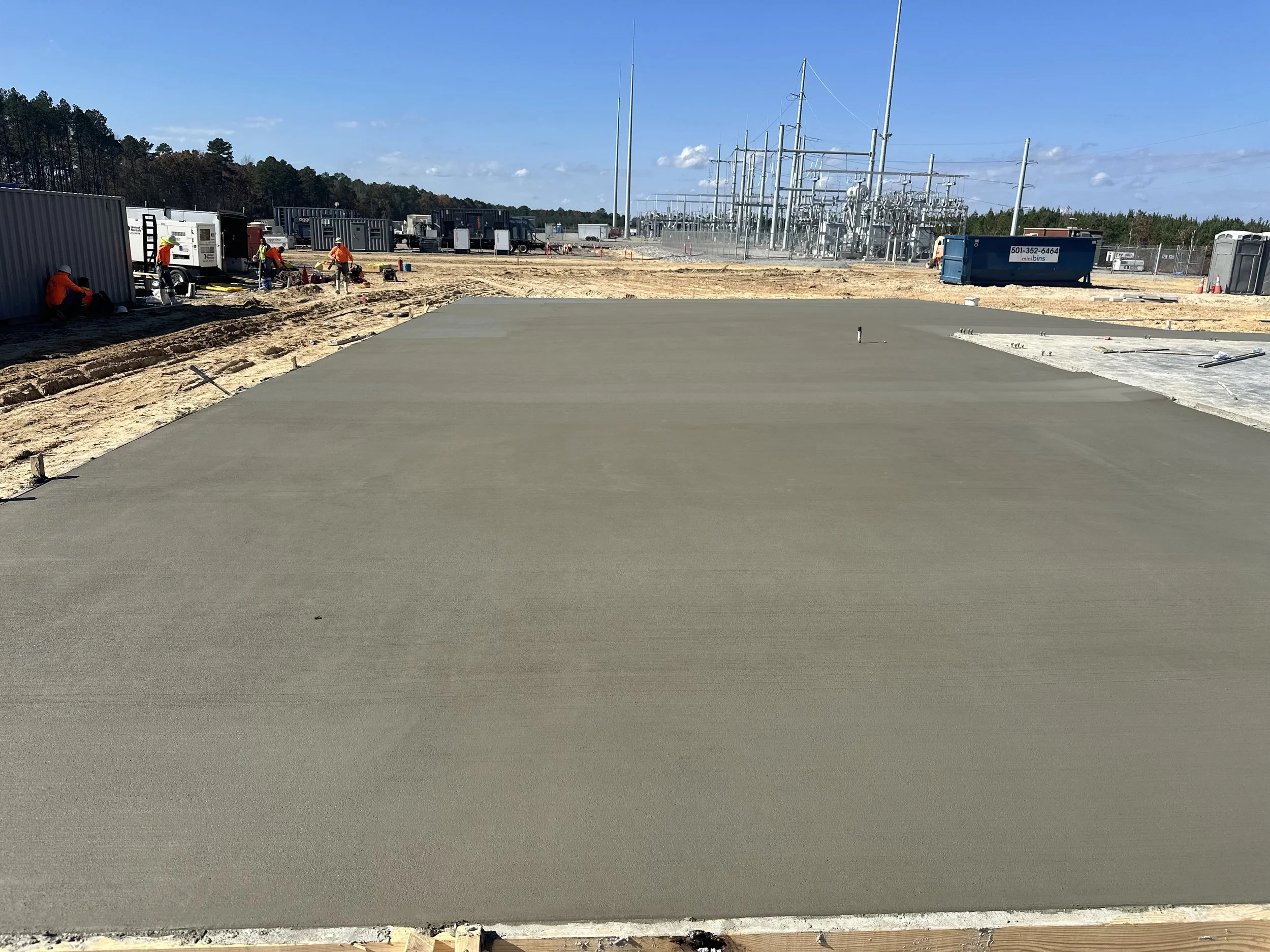 Construction workers pouring and finishing a concrete slab at a construction site with electrical infrastructure in the background.
