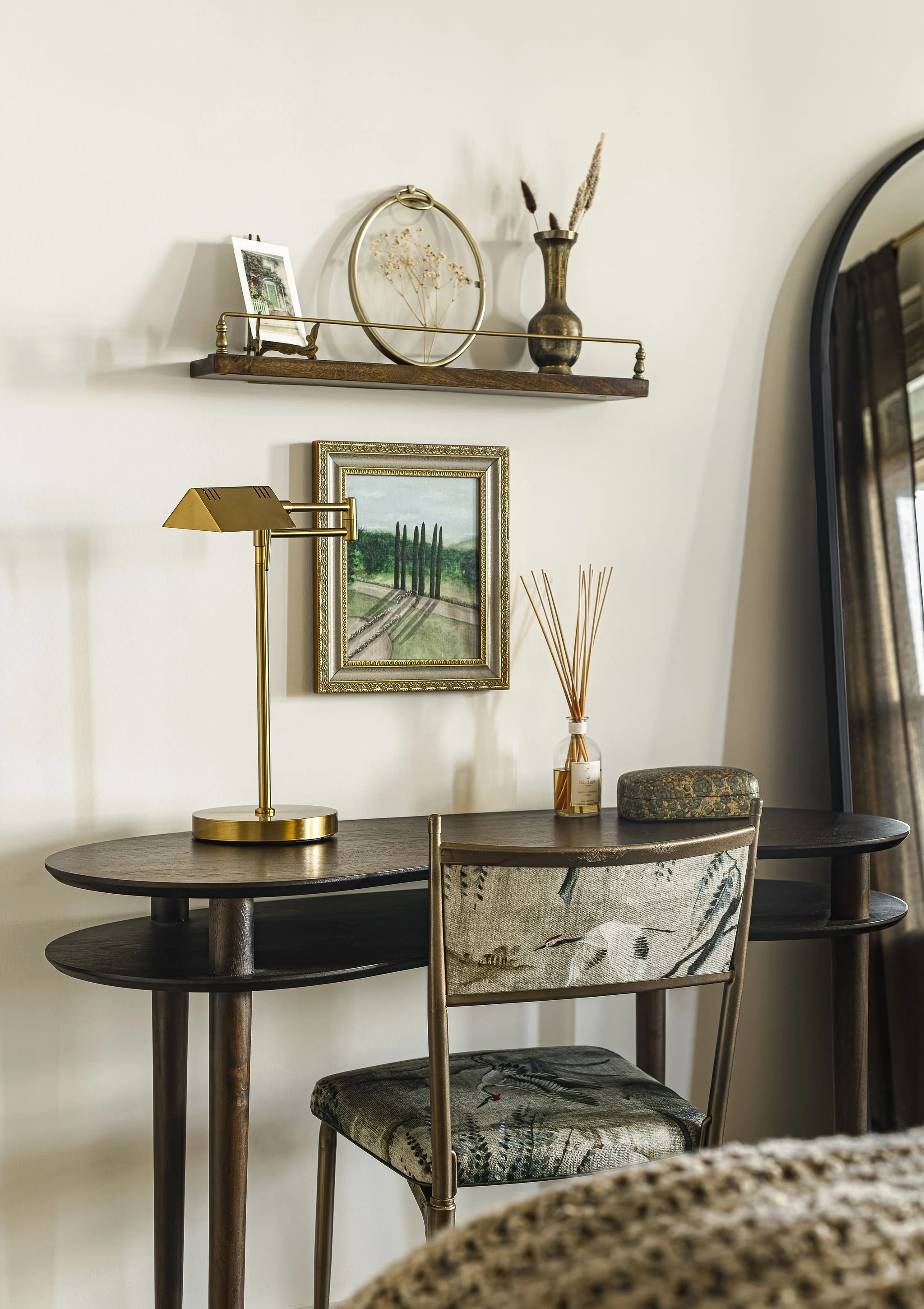 A cozy home office corner with a dark wooden desk, a gold desk lamp, framed landscape art, a glass jar with reed diffusers, a decorative box, and a chair with a bird illustration on the cushion. On the wall, there is a wood and metal shelf with decorative items, plants, and a small photo frame. A large mirror is partially visible on the right.