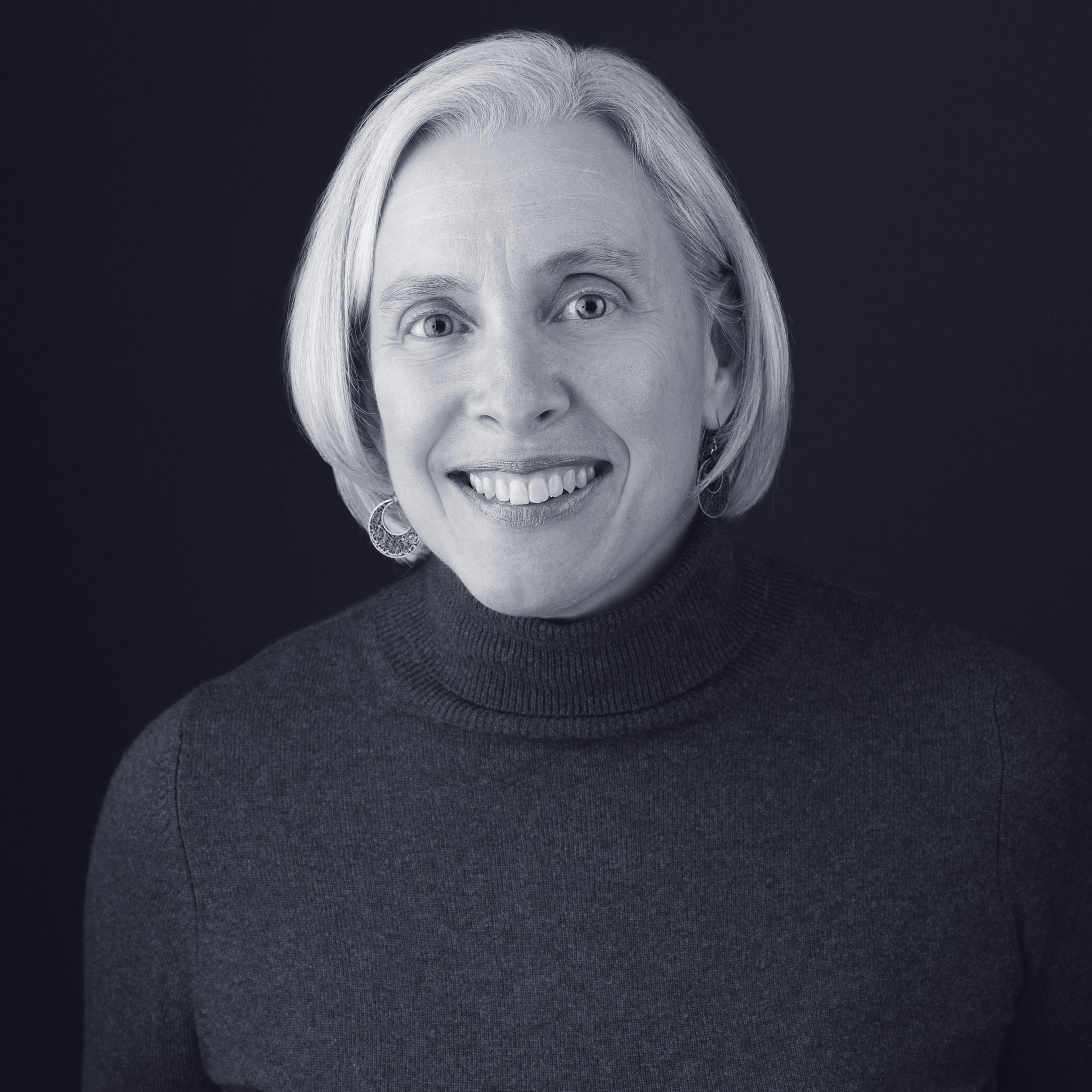 Black and white portrait of a smiling woman with short, light-colored hair wearing a turtleneck sweater and earrings.