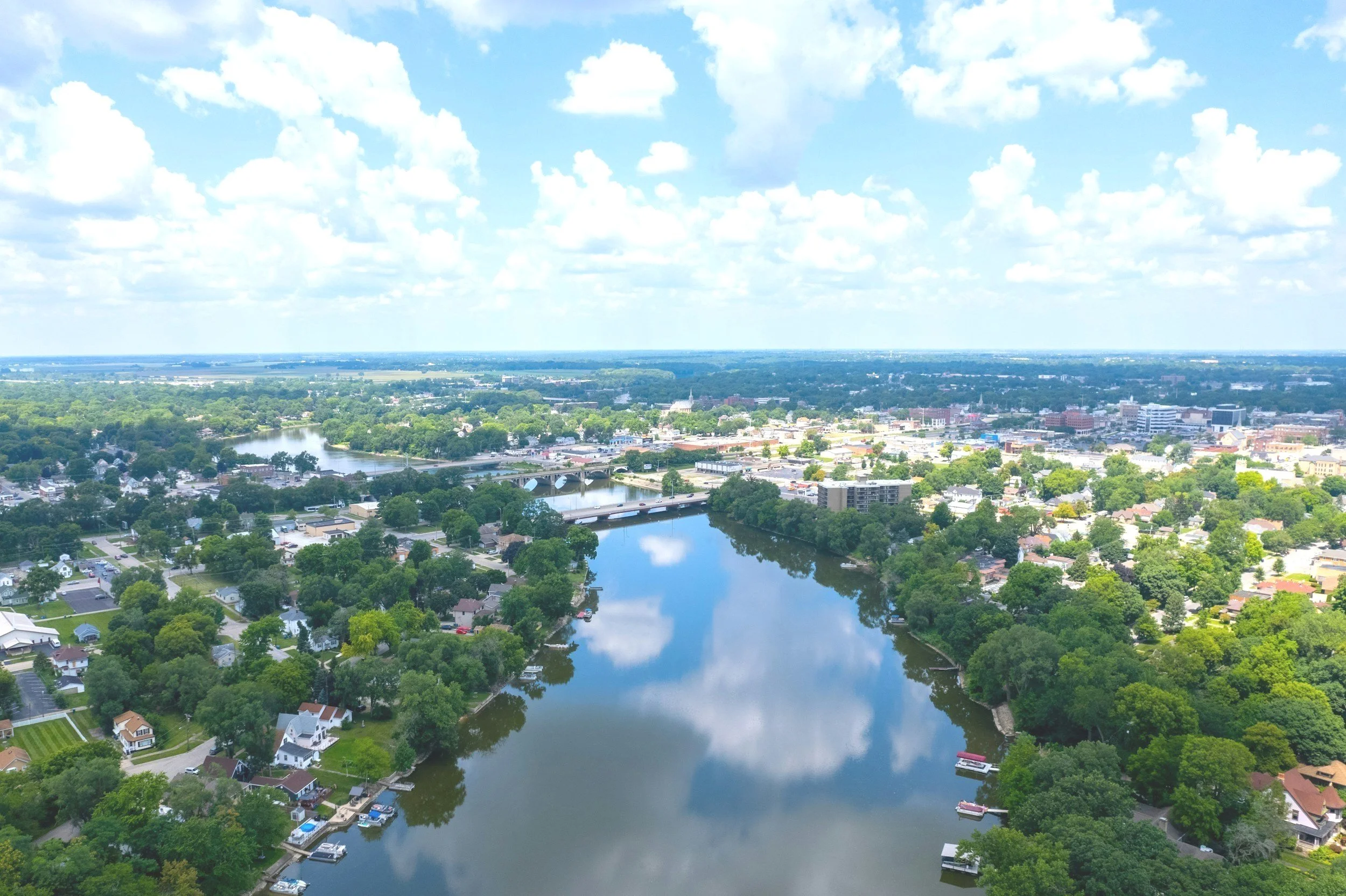 Aerial view of a city with a river running through it, surrounded by green trees and residential houses, under a partly cloudy sky.