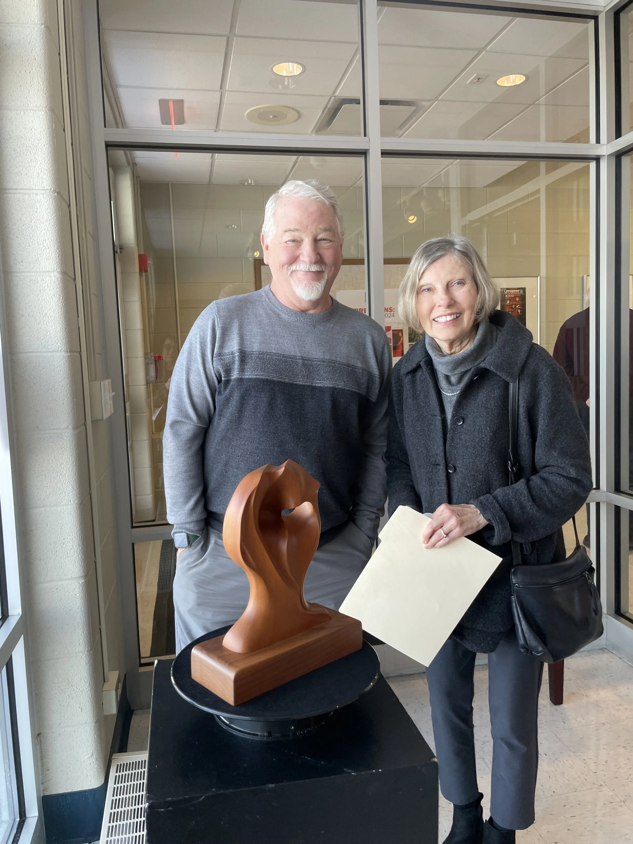A smiling elderly man and woman standing next to a wooden abstract sculpture in an indoor setting.