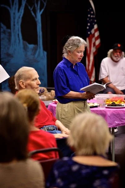 An elderly woman in a blue shirt reading a book to a group of people at a gathering, with an American flag in the background and a table covered with a pink tablecloth.