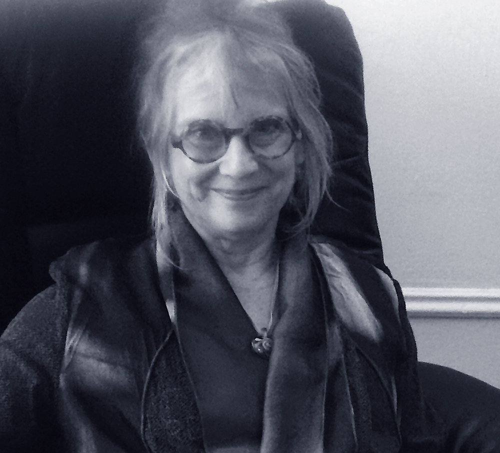 A woman with glasses and shoulder-length hair smiling, sitting in a black leather chair, wearing a dark shirt with a scarf, in black and white.