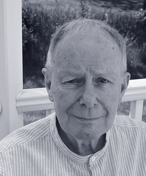 A grayscale photo of an elderly man with short light hair, wearing a striped shirt, sitting outdoors in front of a window with a landscape background.