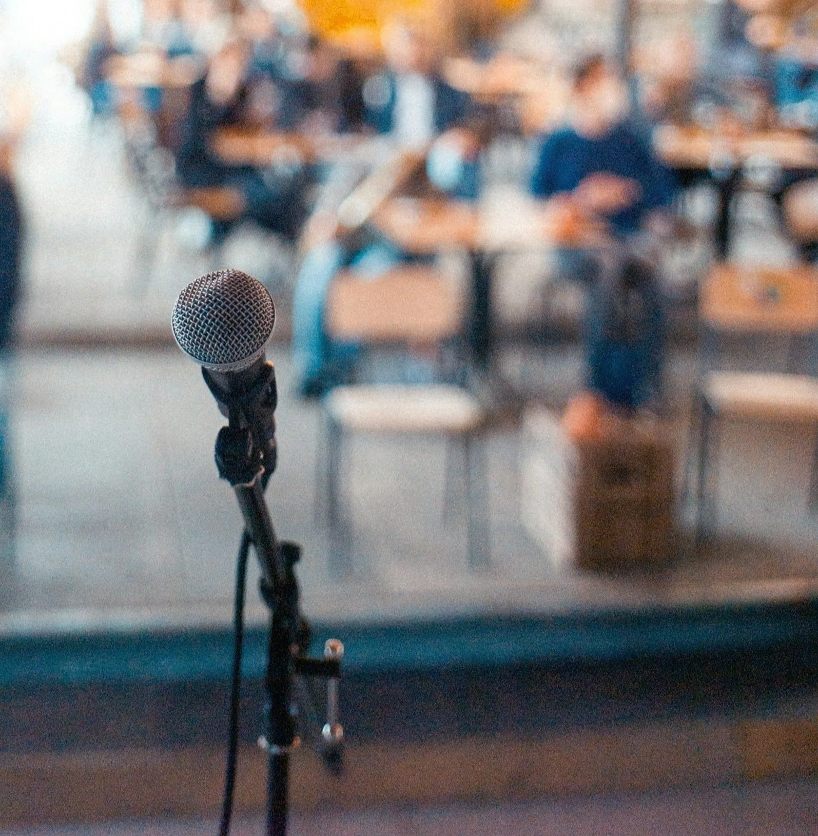 A microphone on a stand facing an audience in a blurred background, suggesting a stage or presentation setting.