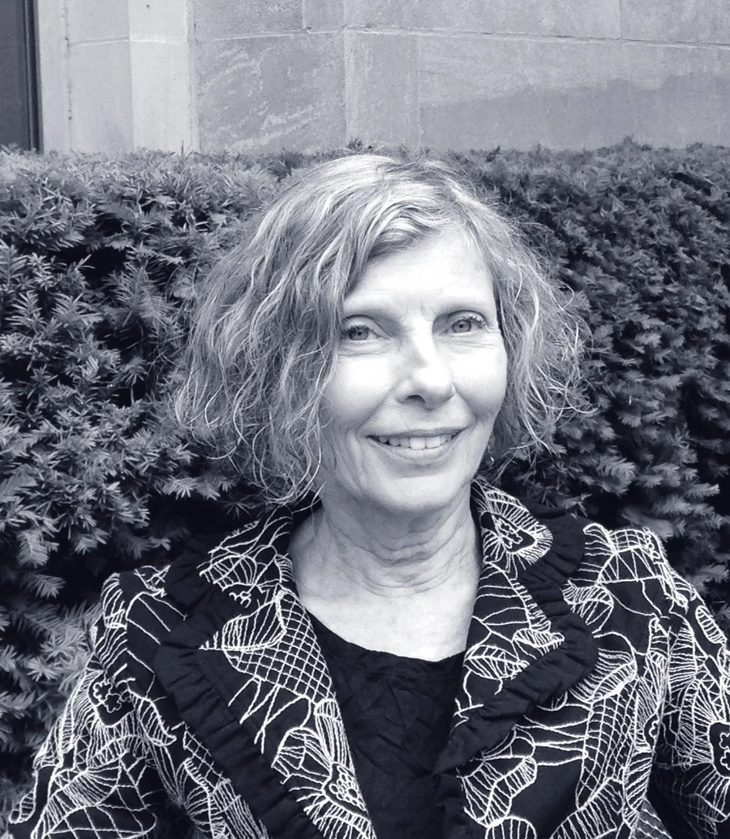 A middle-aged woman with curly hair smiling outdoors, sitting in front of a bush and a building wall.