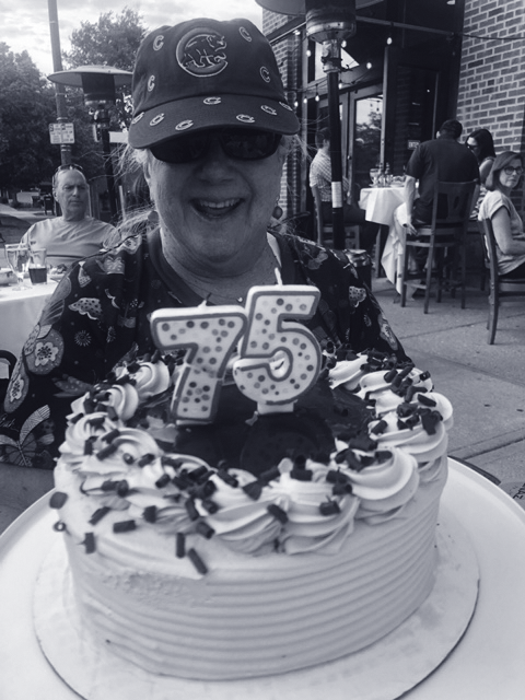 A woman smiling at a 75th birthday celebration outdoors with a decorated cake featuring a large "75" candle and black and white icing and sprinkles.