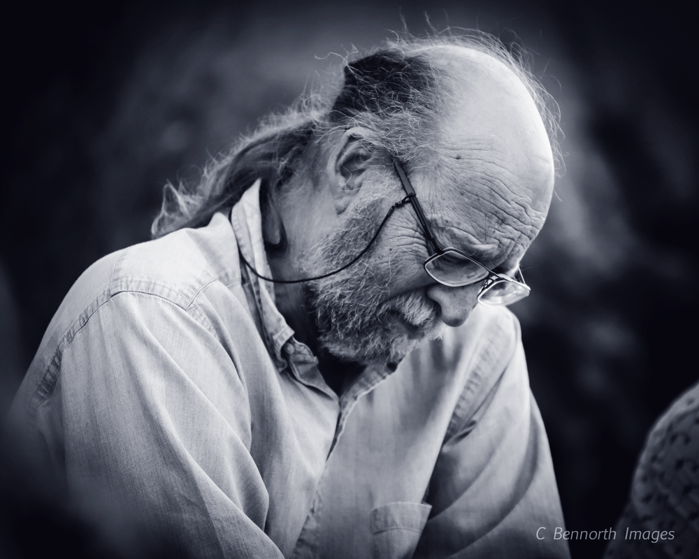 An elderly man with long gray hair and beard, wearing glasses, looking downward in a moment of reflection.