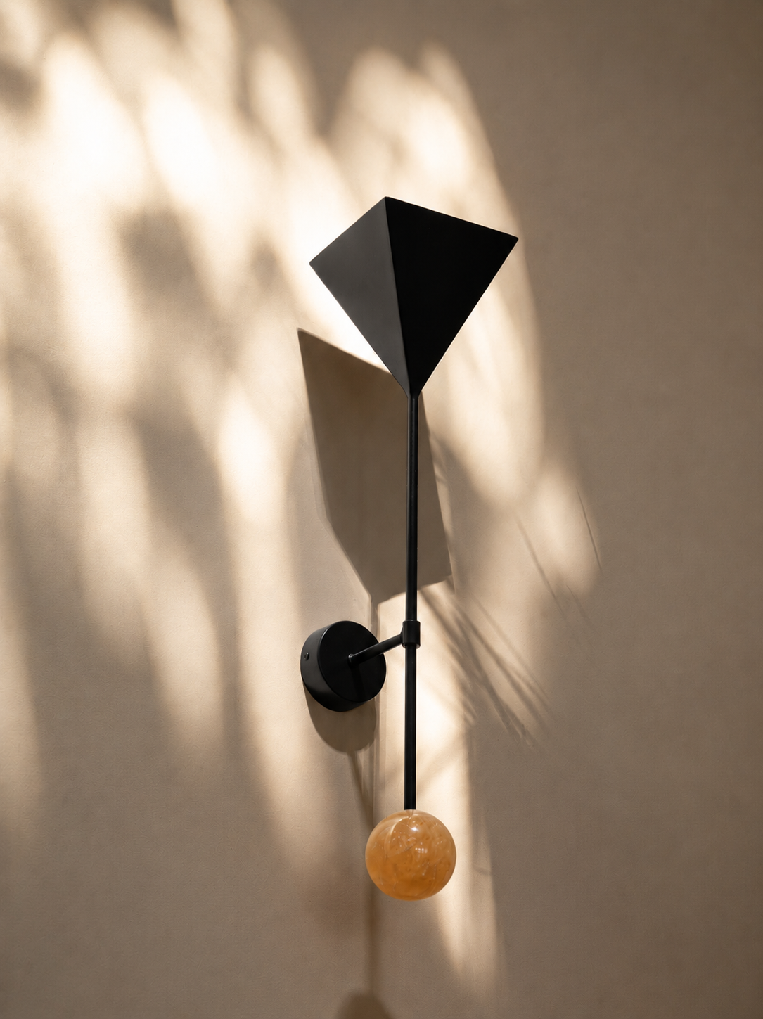 Modern wall sconce with black geometric shade, black rod, and orange marble sphere, casting decorative shadows on beige wall.