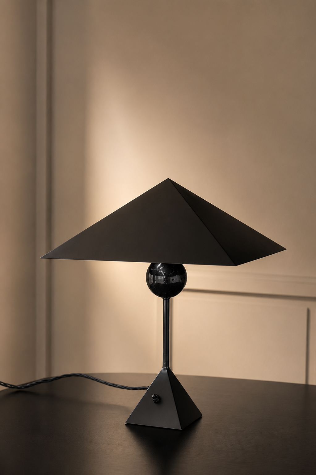 Black modern table lamp with a geometric design and a black sphere at its center, placed on a dark tabletop in a dimly lit room.
