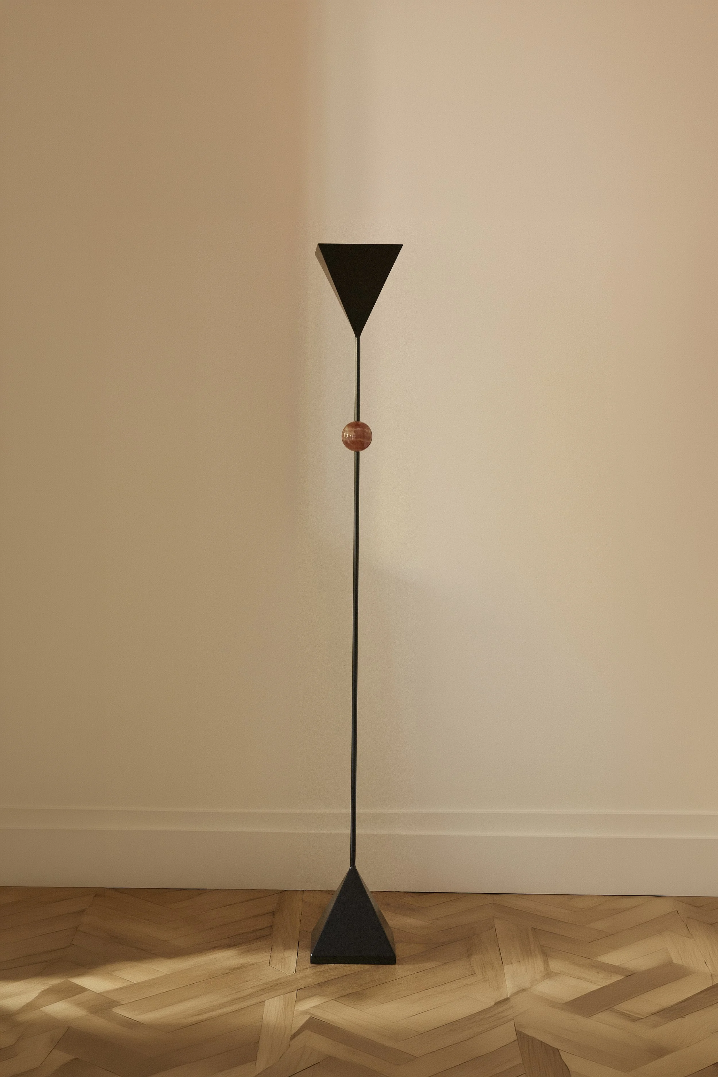 A modern floor lamp with a black base, a thin metal pole, and a black cone-shaped shade. The lamp has a decorative spherical element near the top of the pole. It stands on a wooden parquet floor against a plain wall.
