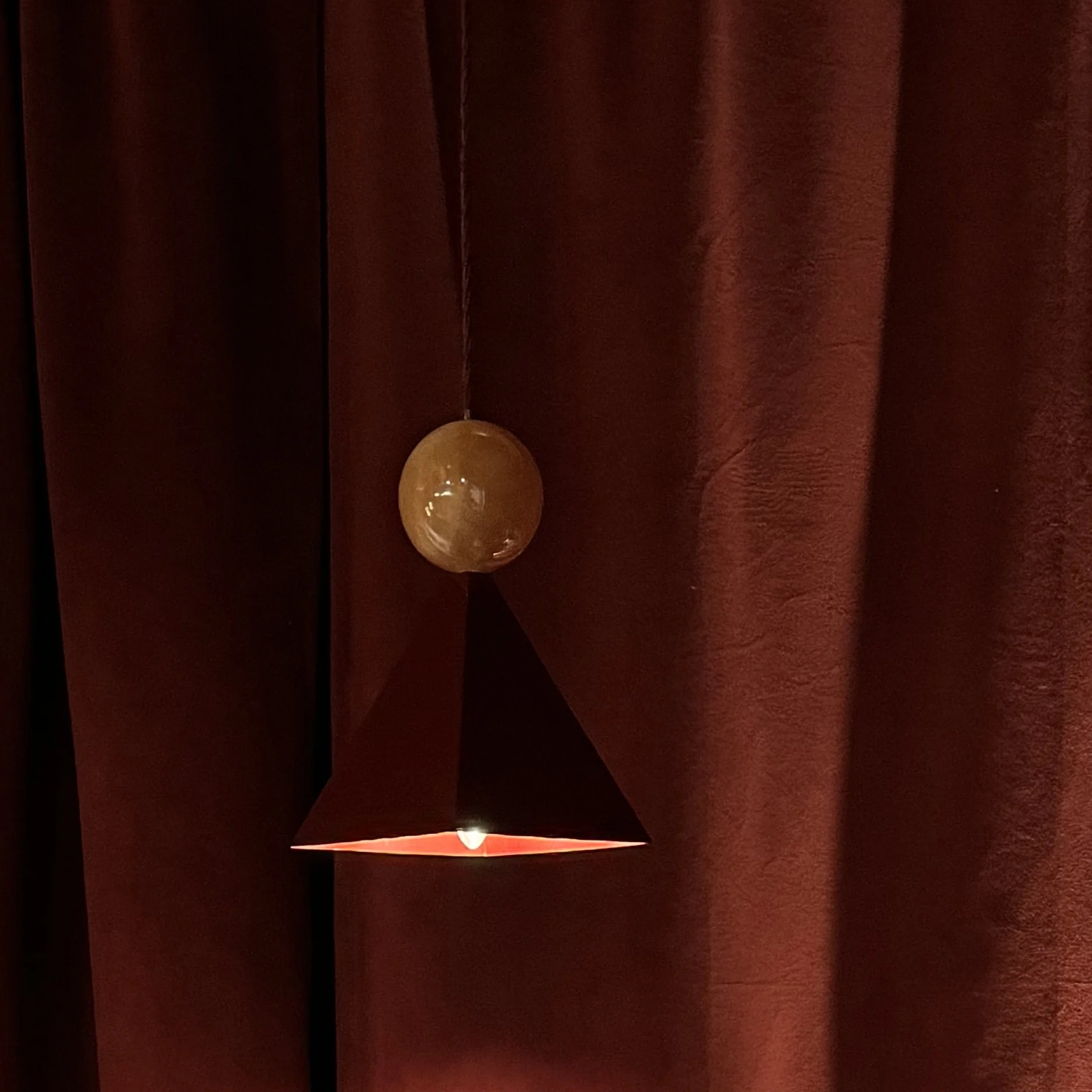 Close-up of a modern pendant light with a glowing bulb, hanging in front of a dark red curtain.