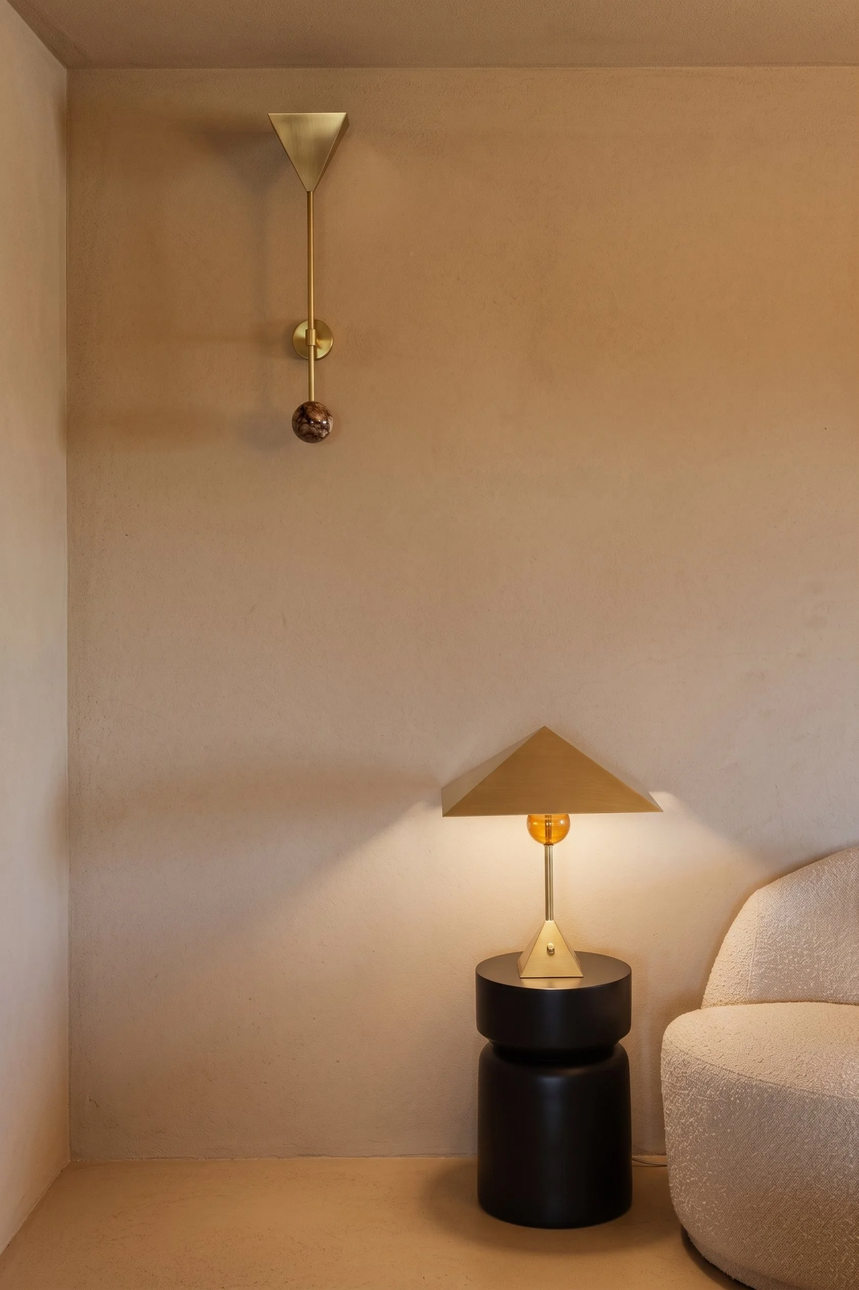 A beige textured wall with a gold wall sconce and a table lamp with a gold pyramid-shaped shade, black base, and orange bulb, next to a rounded beige armchair.