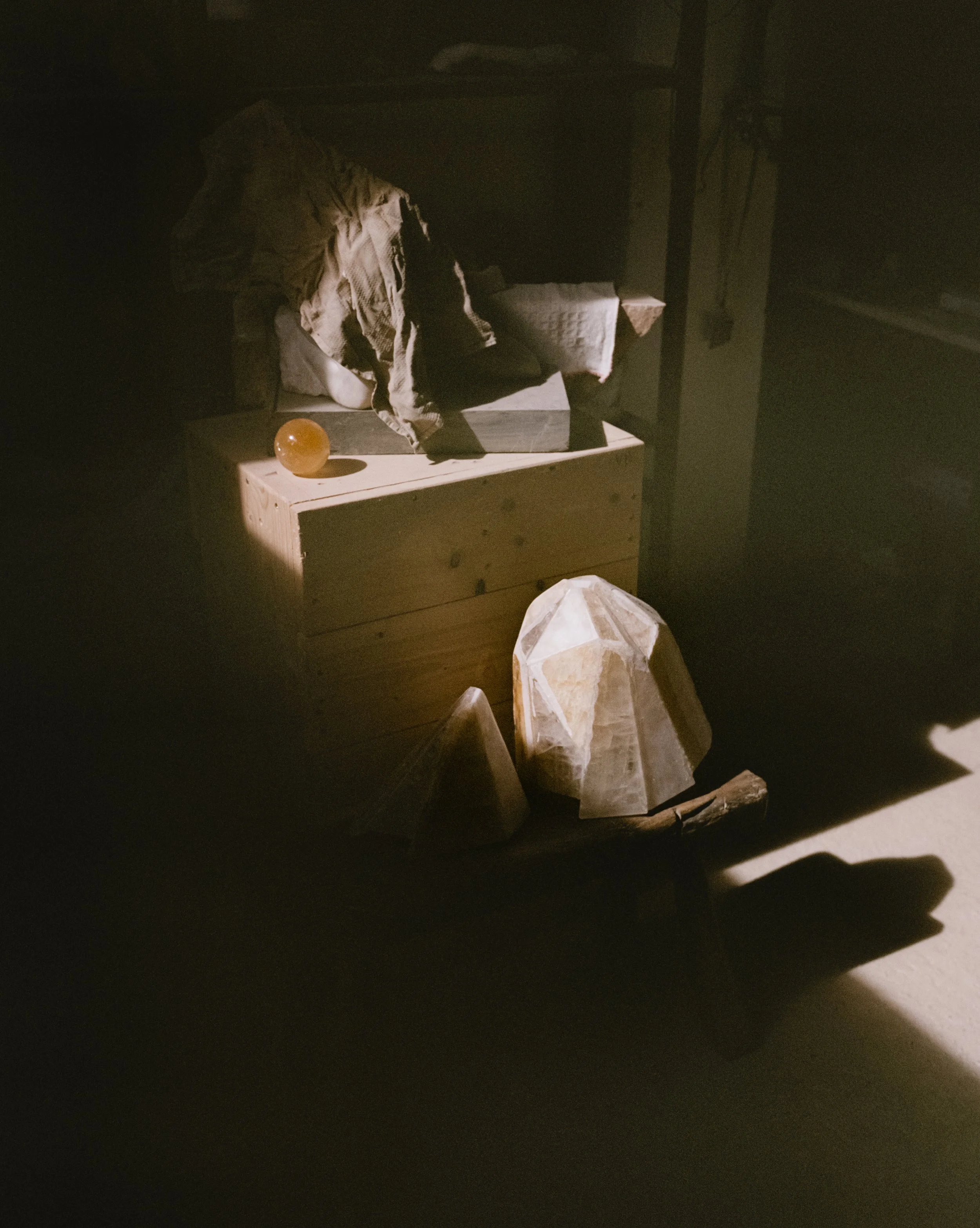 A dark room with a wooden table holding a translucent orange sphere and some crumpled paper. There are large rocks and a geometric stone sculpture on the floor, with a minimalist shelving unit and a fabric-covered surface in the background.