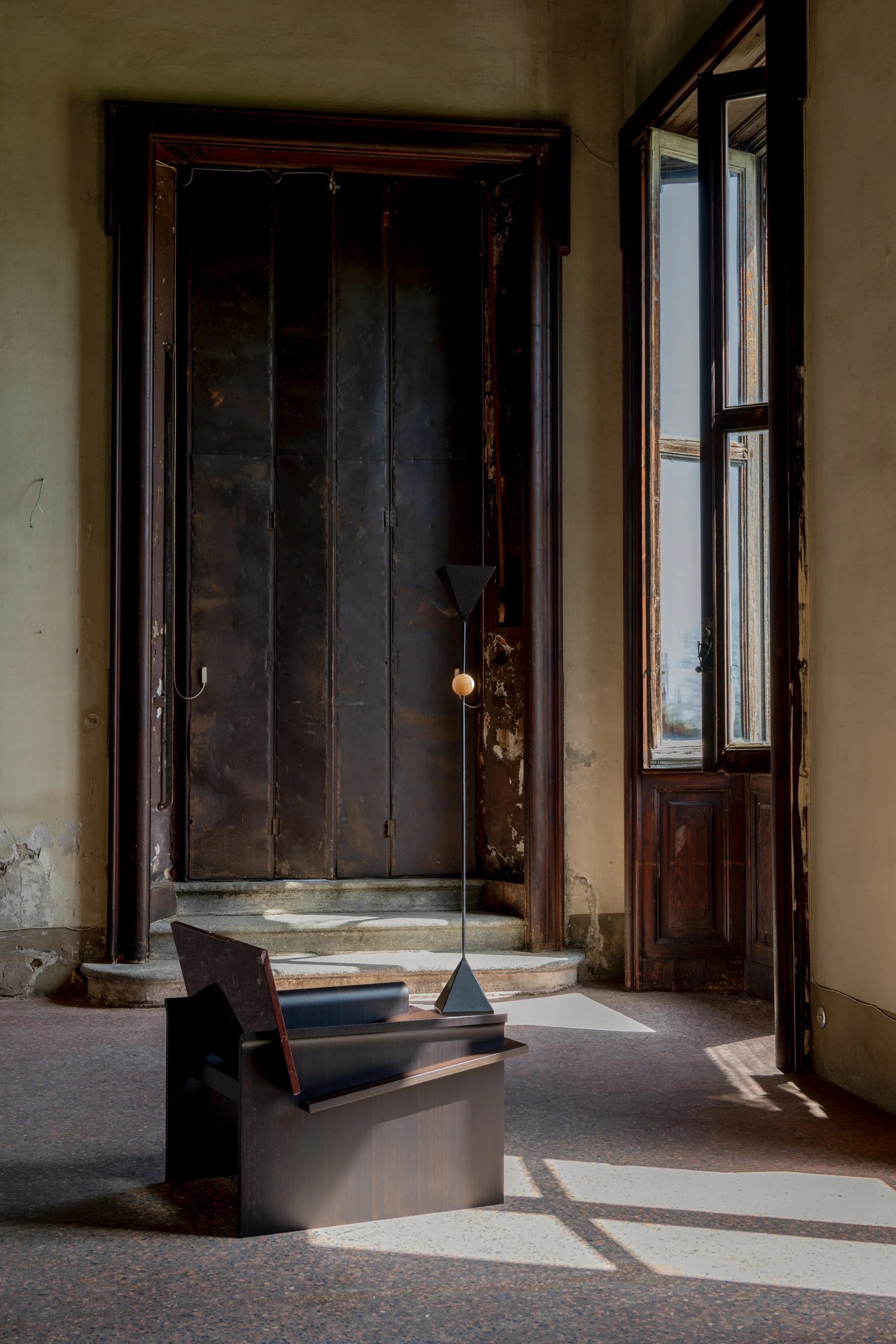 Interior of an old building with large open wooden windows, an antique metal wardrobe or cabinet, a modern sculpture, a bench, and sunlight casting shadows on the floor.