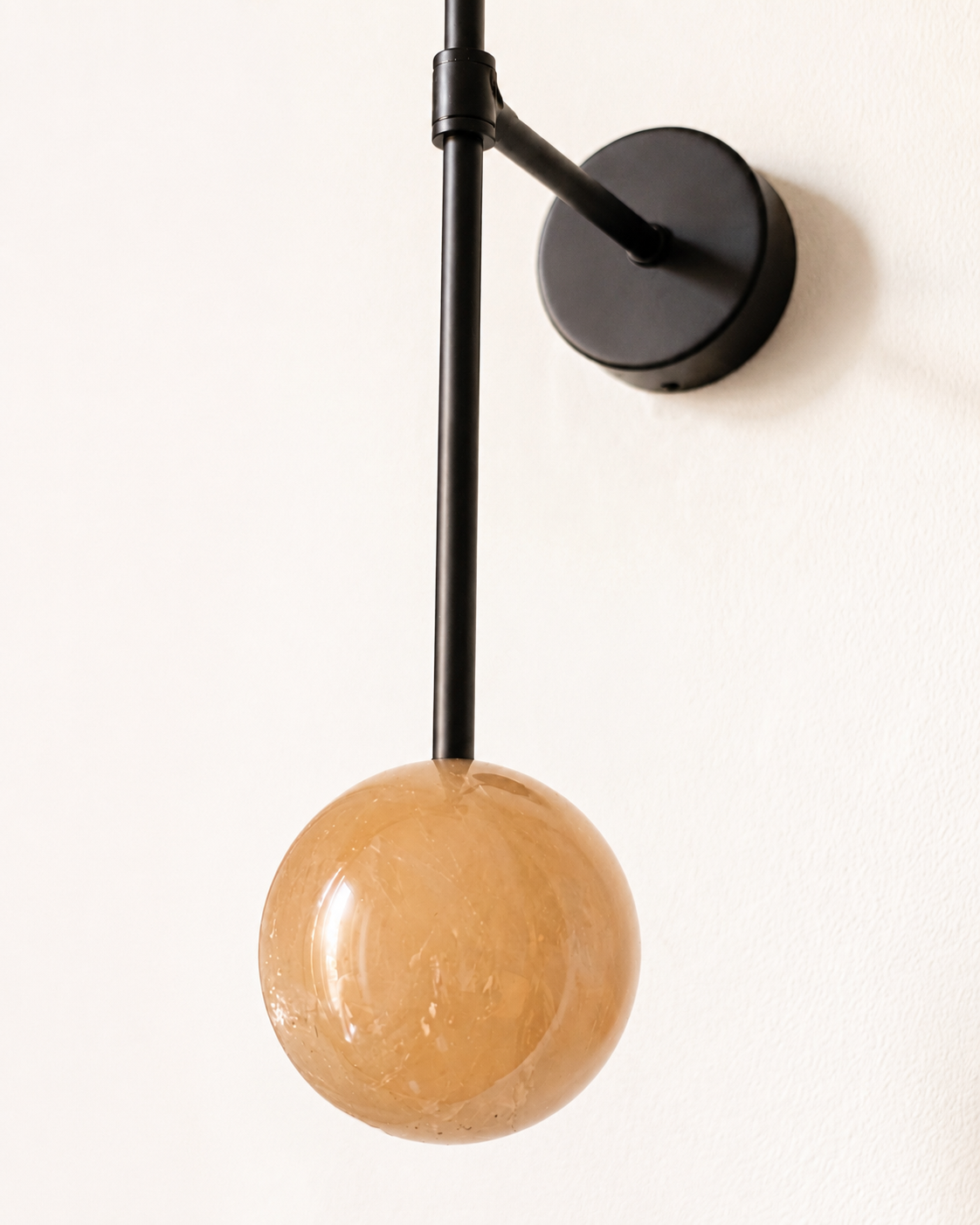 A modern wall-mounted light fixture with black metal arm and spherical amber glass shade, attached to a white wall.
