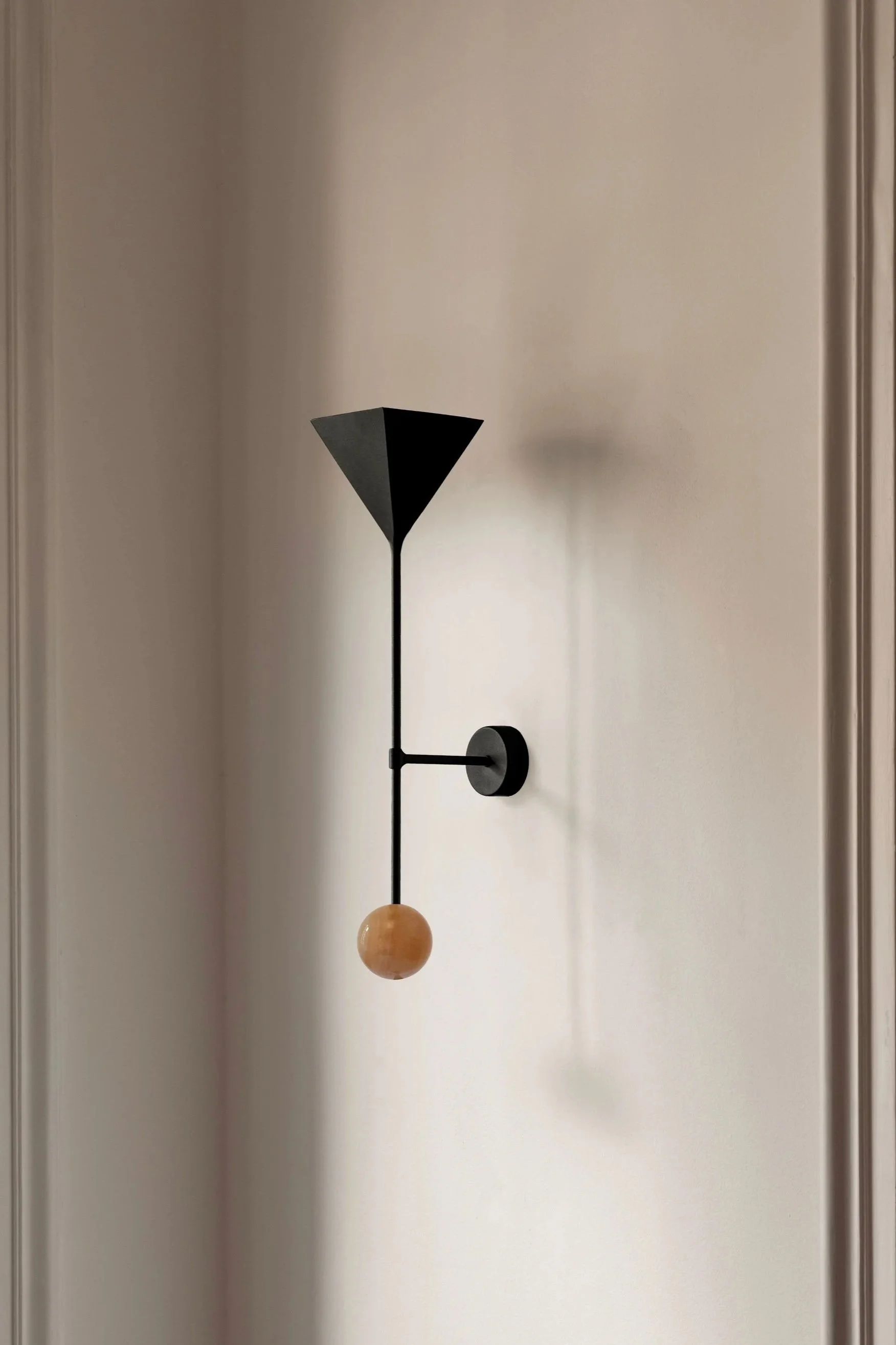 A black and beige wall-mounted mobile with a black cone-shaped element, a vertical black rod, a horizontal black bar, and a wooden sphere at the bottom, hanging on a white wall.