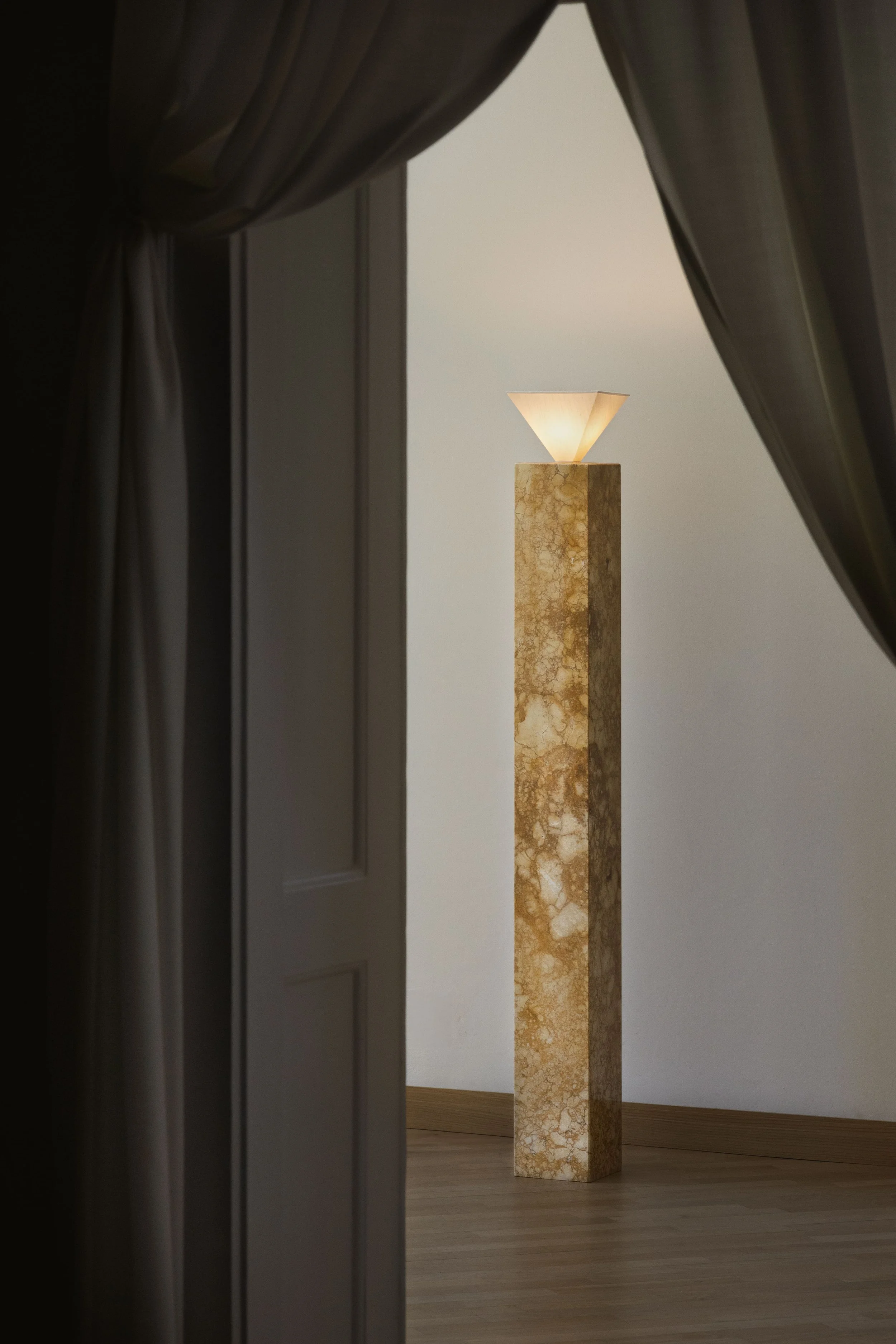 A lamp with a beige and brown marble base and a conical beige lampshade is partially visible through a dark curtain and door frame, standing on a wooden floor against a plain light-colored wall.