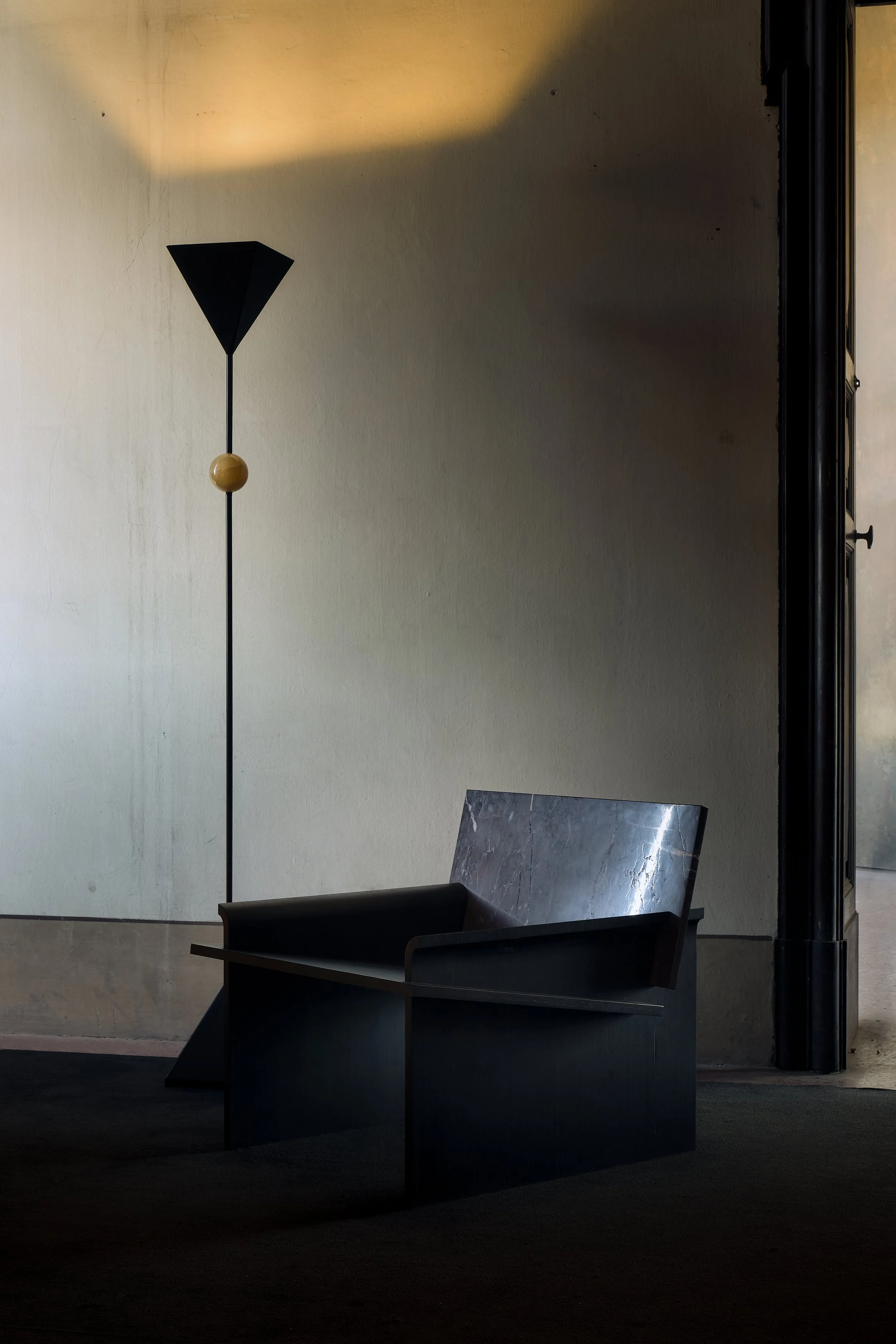 Interior view of a modern waiting area with a black bench and a distinctive floor lamp with a spherical detail, against a plain wall with subtle lighting and shadows.