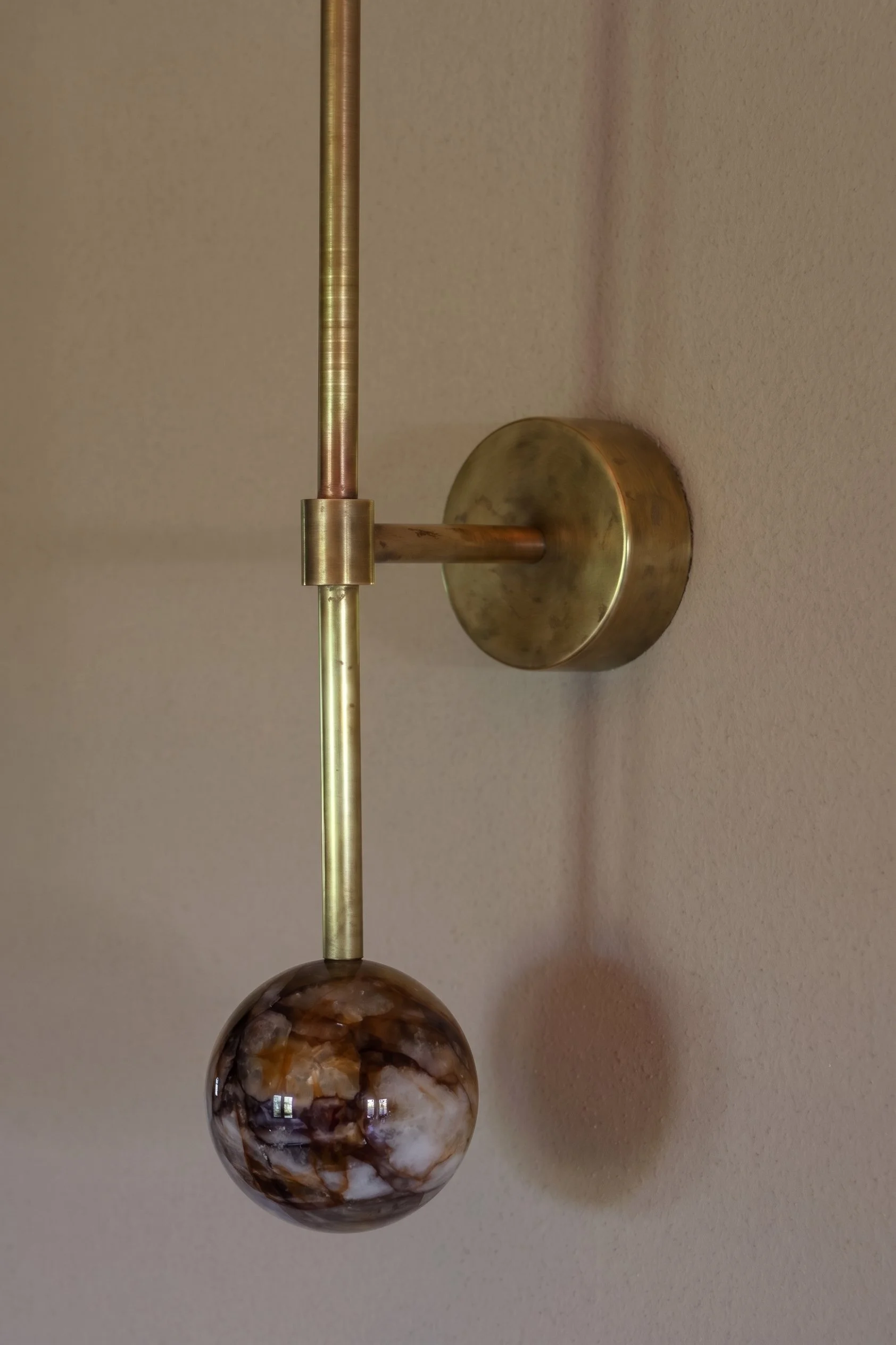 Close-up of a modern wall-mounted lamp with a brass finish and a marble sphere at the bottom, against a beige textured wall.