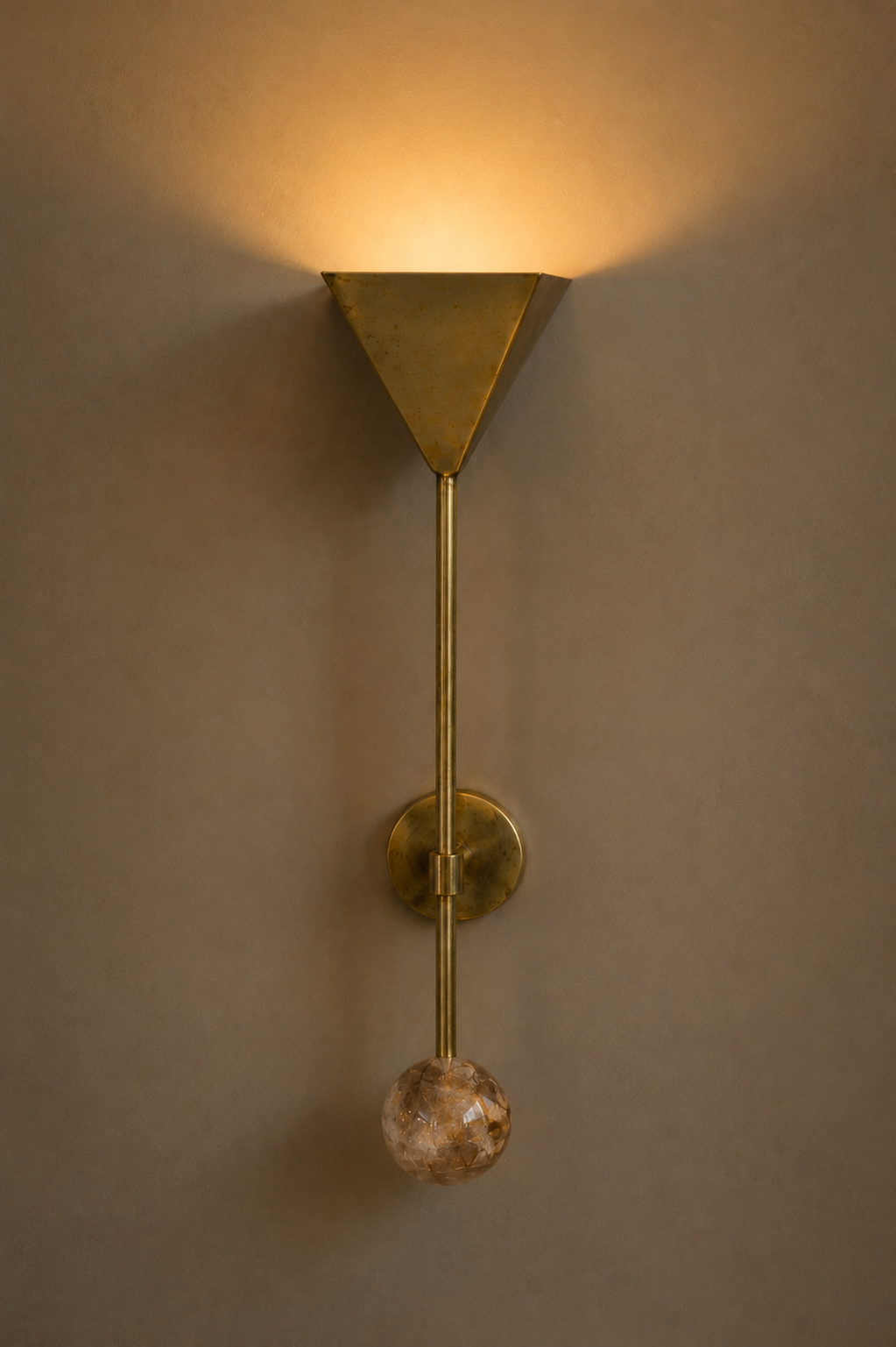 A modern wall-mounted light fixture with a brass finish, featuring a trapezoidal light cover and a glass ball at the bottom, mounted on a beige wall.