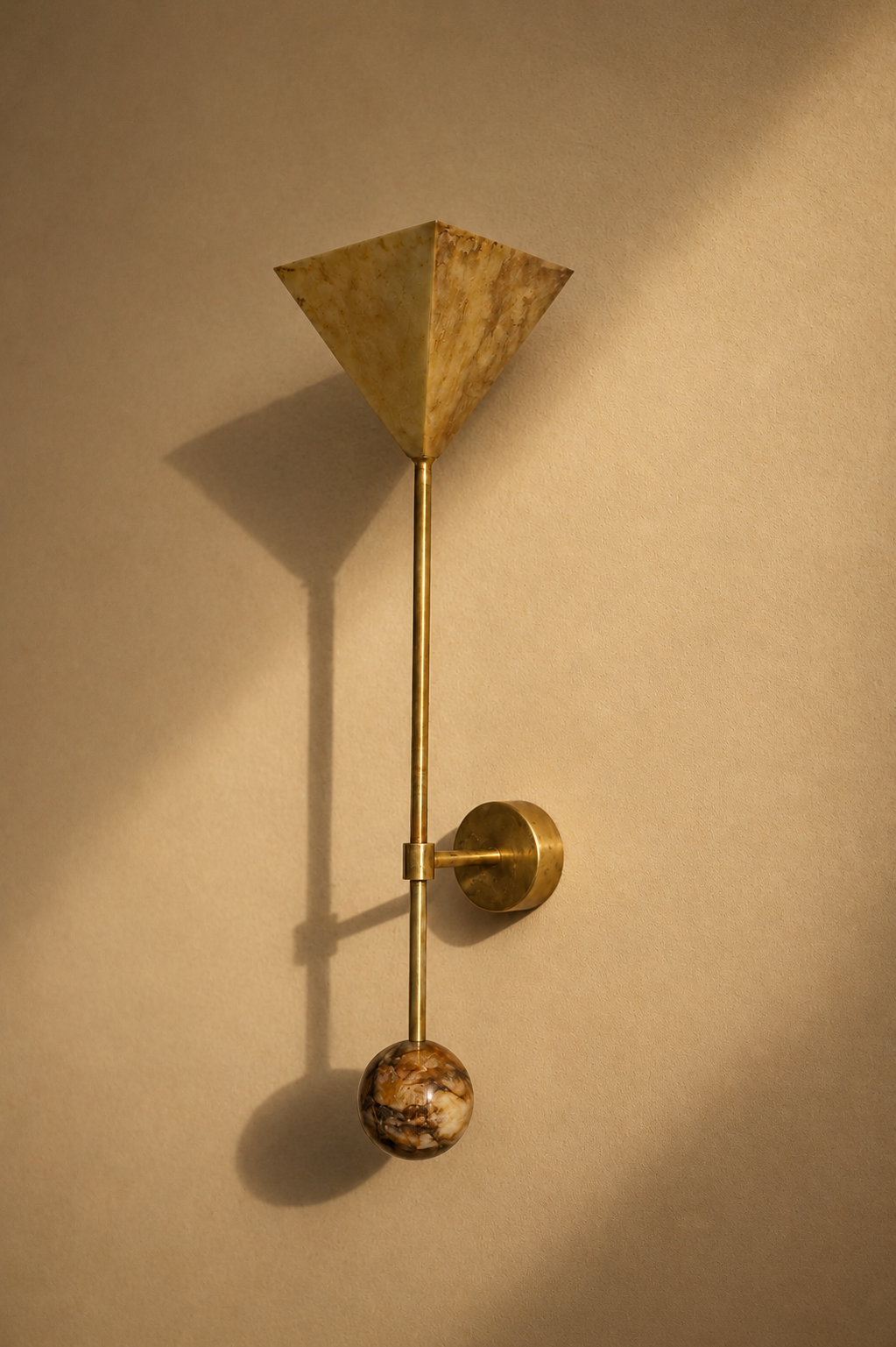 A modern wall-mounted lamp with a brass finish, featuring a geometric marble shade at the top, a slim vertical brass rod, a circular brass fixture, and a marble sphere at the bottom, casting shadows on a beige wall.
