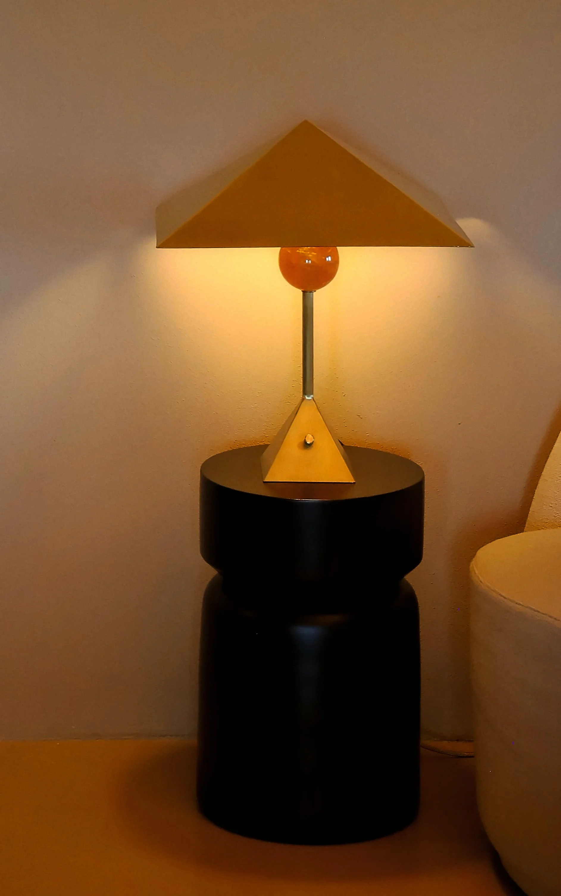 A table lamp with a yellow conical shade, a yellow triangular base, and a yellow spherical connector, placed on a black round side table in a dimly lit room.