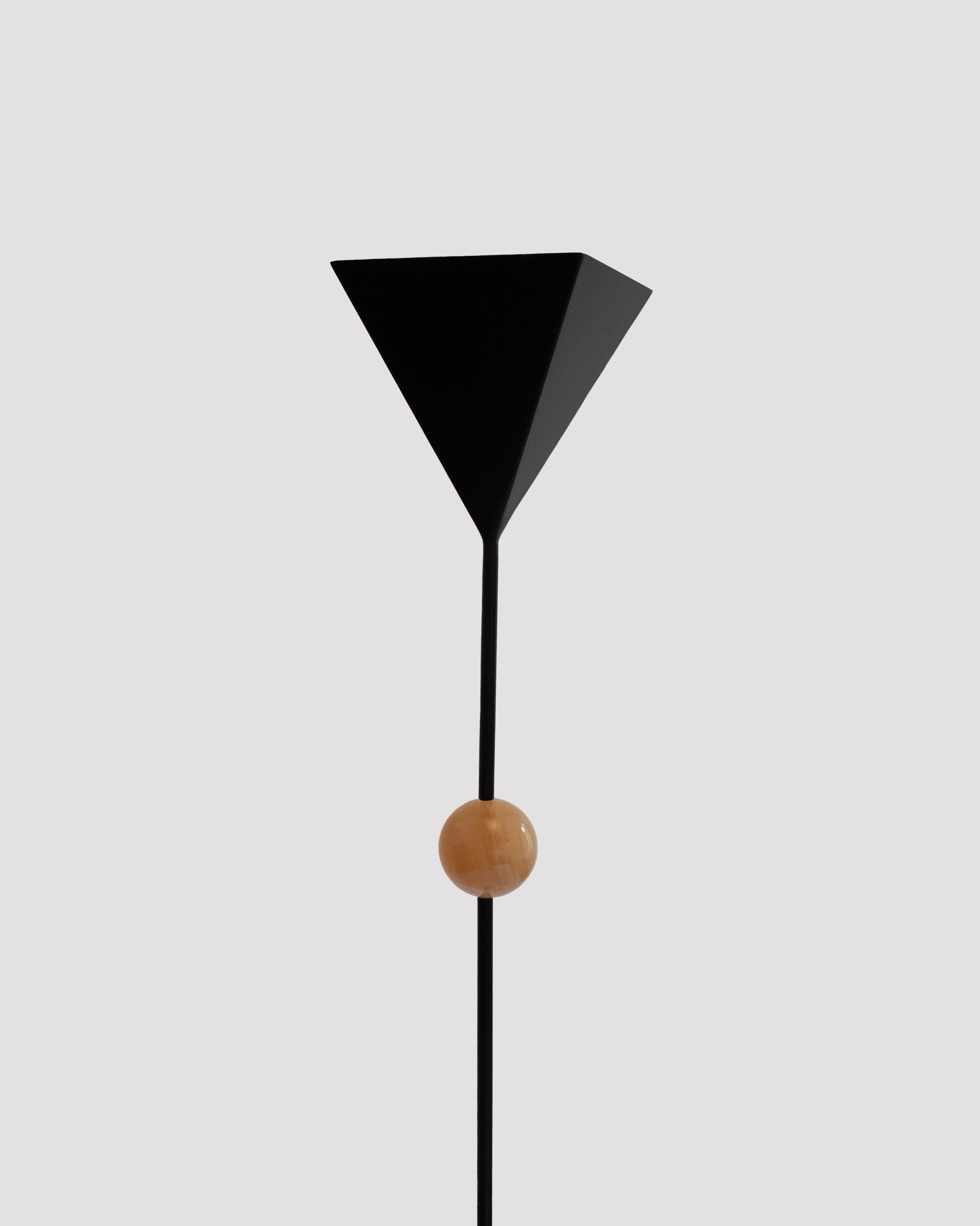 A black, modern sculpture on a black metal stand featuring a large black geometric shape at the top, a round beige element in the middle, and a smaller black element at the bottom, set against a plain light gray background.