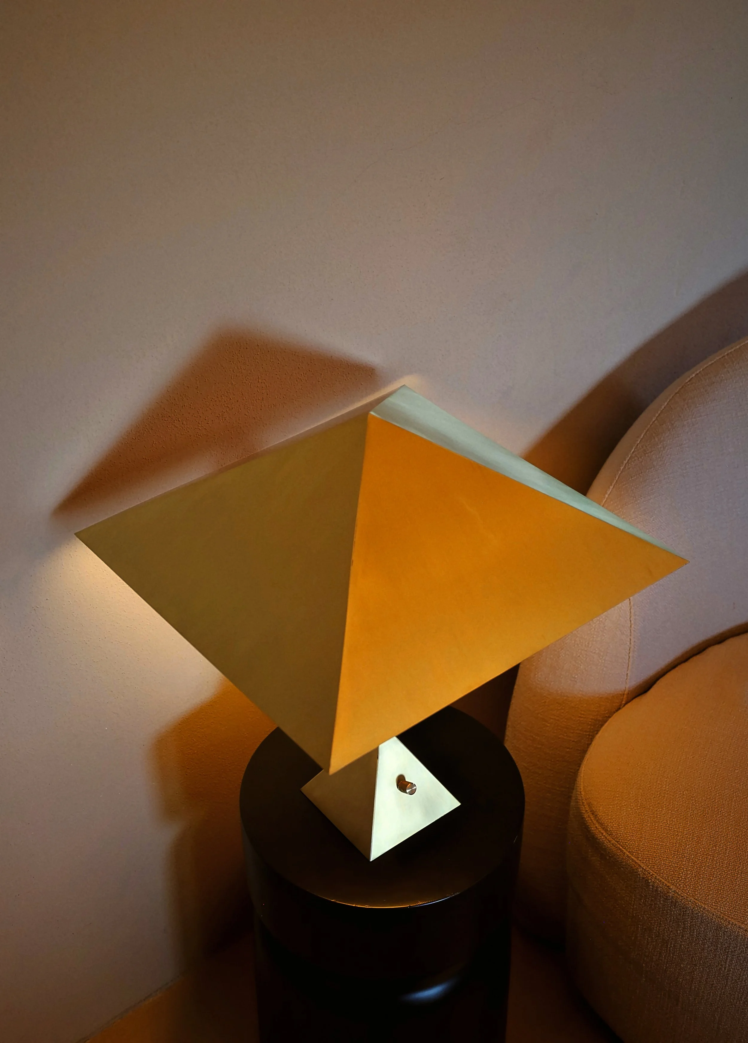 A modern table lamp with a gold-colored, angular shade and a black circular base, placed on a small round black side table next to a beige upholstered chair.