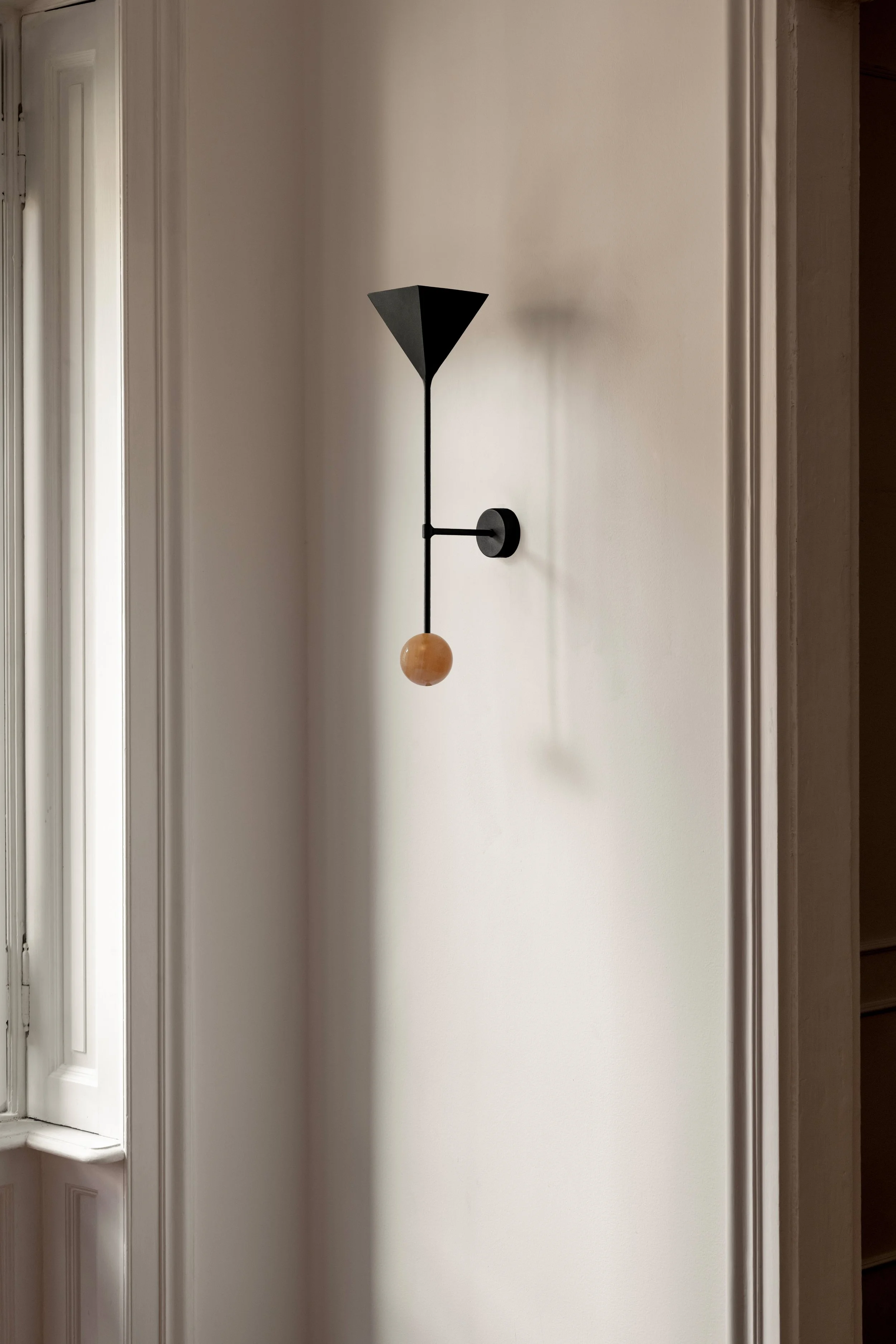 Modern wall sconce with black geometric lampshade and wooden ball detail, mounted on a plain white wall near a window.