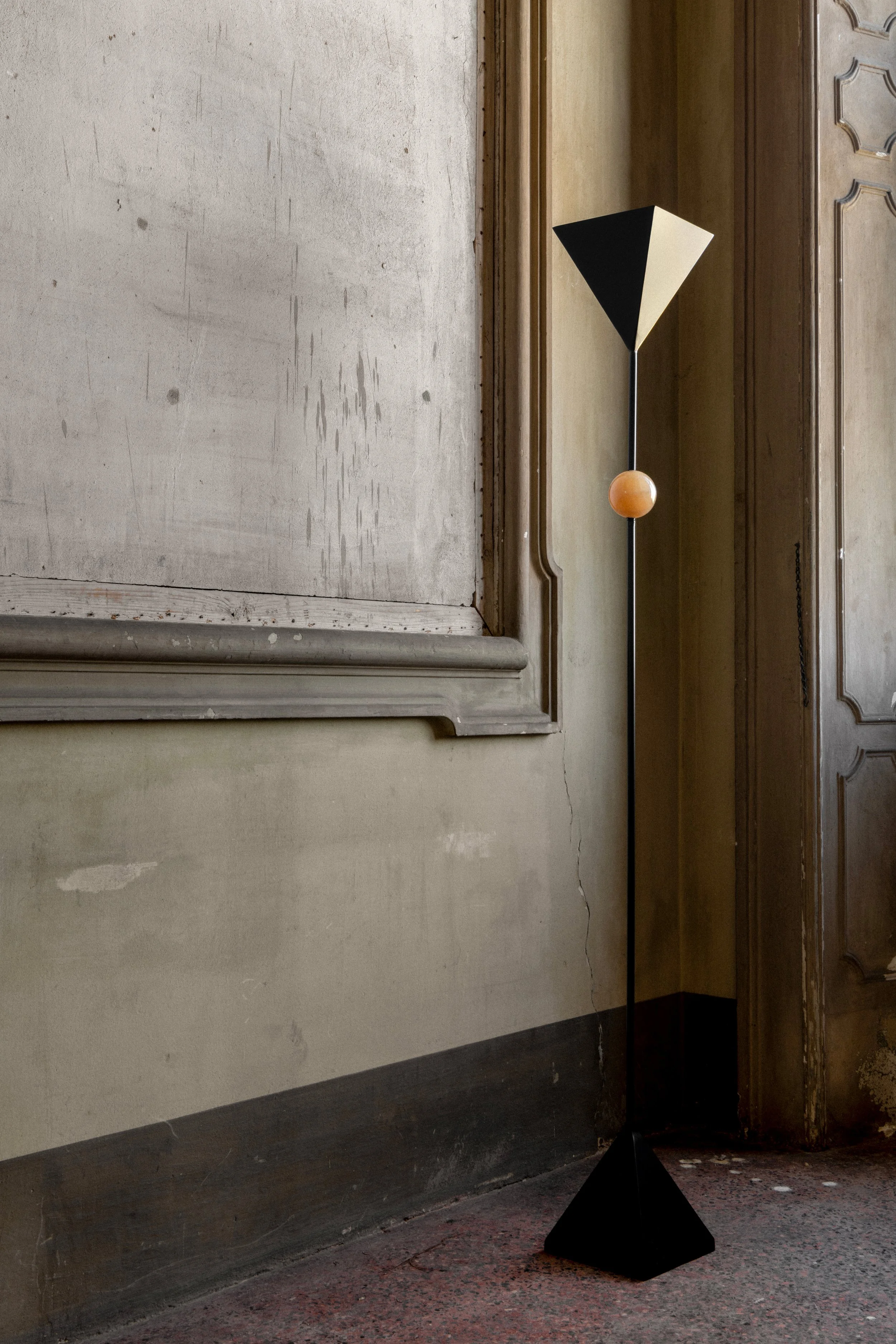A modern floor lamp with a geometric black and beige shade and a spherical wooden accent at the base, standing against a vintage wooden paneled wall in a room with a concrete wall and a granite floor.