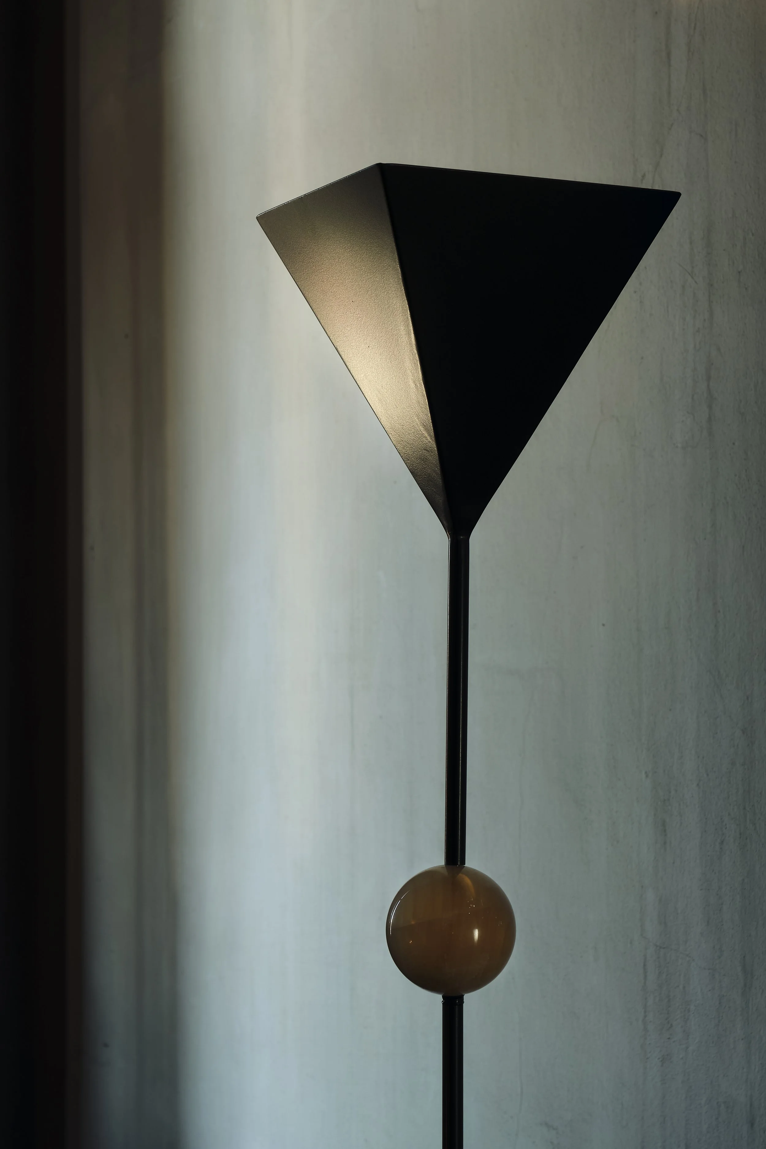 Modern floor lamp with a black, angular shade, a round wooden base, and a thin black stand, positioned against a lightly textured wall with soft natural light.