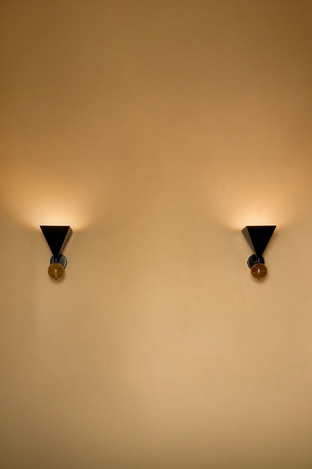 Two wall sconces with black triangular shades and round bulbs, mounted on a beige wall, casting warm light.