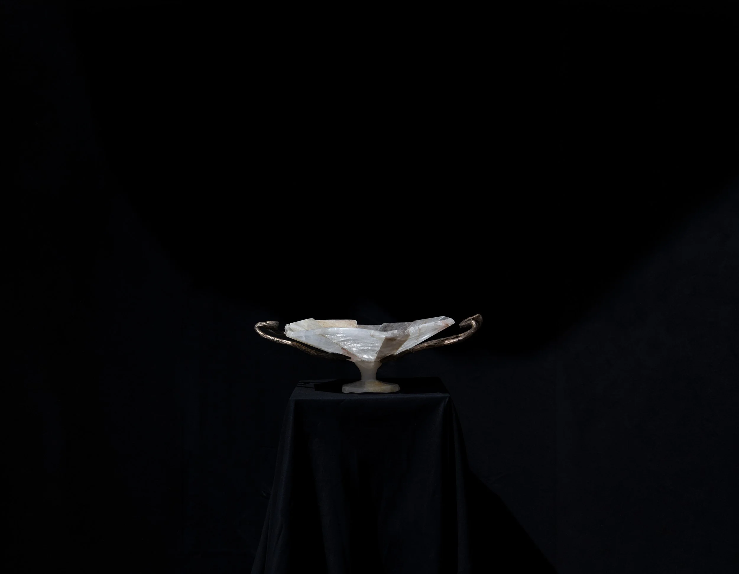 A large, clear, faceted crystal on a decorative stand displayed on a black pedestal against a black background.