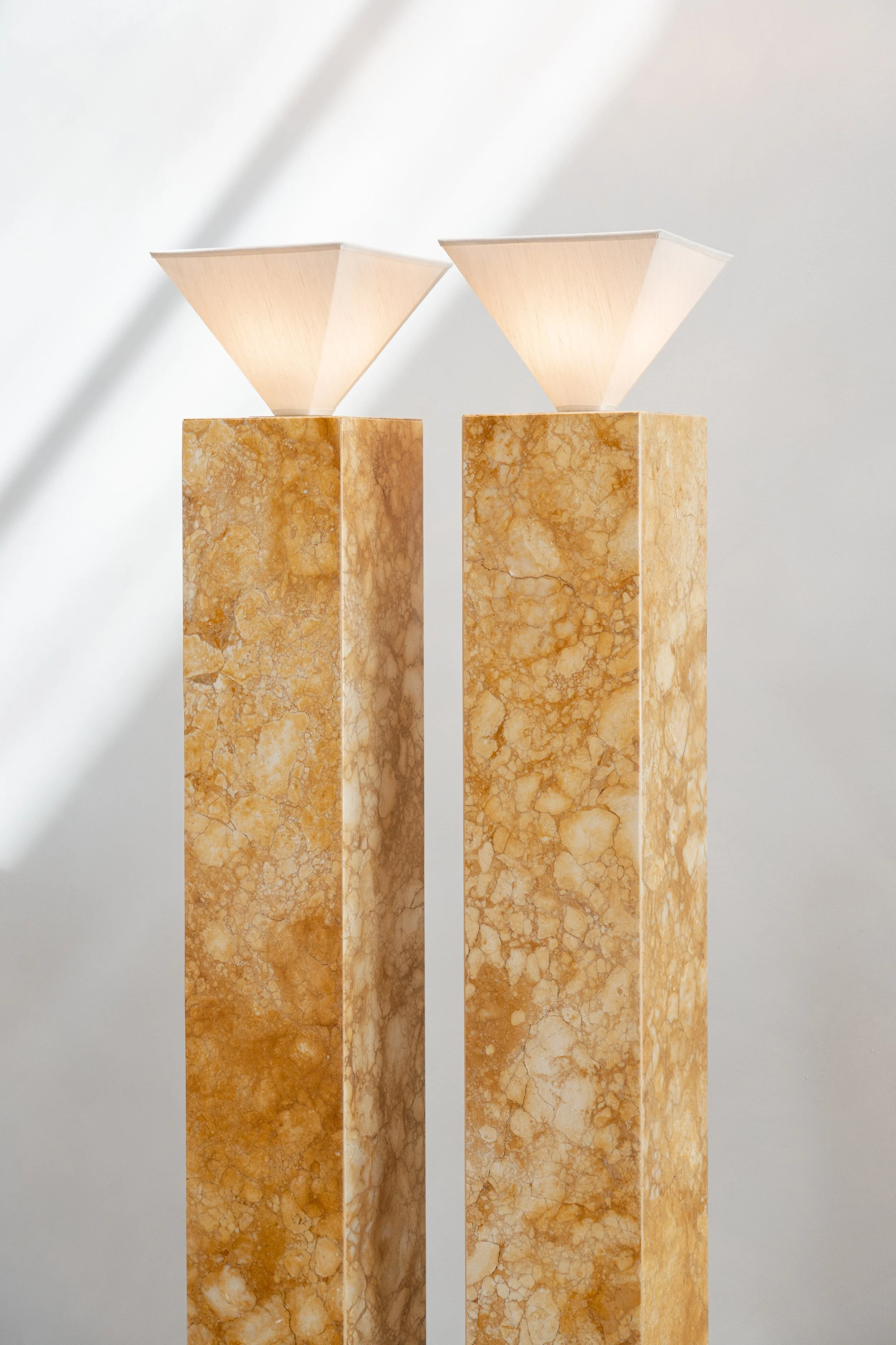 Two tall, beige marble pedestals with white, geometric, cone-shaped lamps on top against a plain, light background.