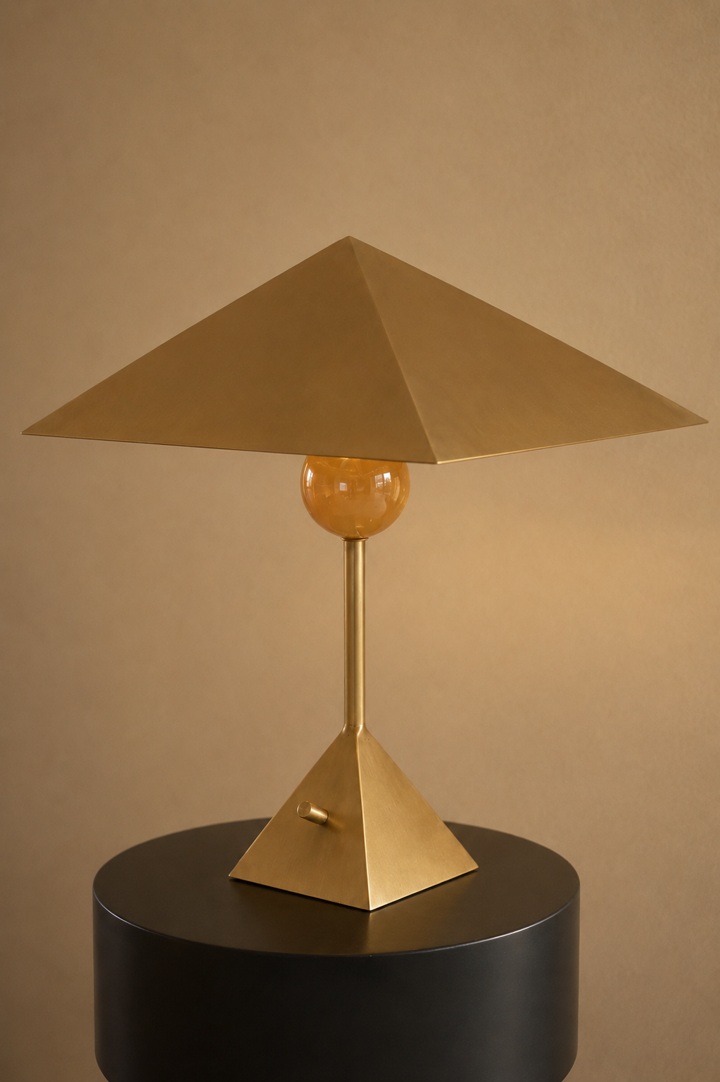 A modern table lamp with a geometric gold base, a spherical accent, and a pyramid-shaped shade, placed on a black cylindrical table against a beige wall.