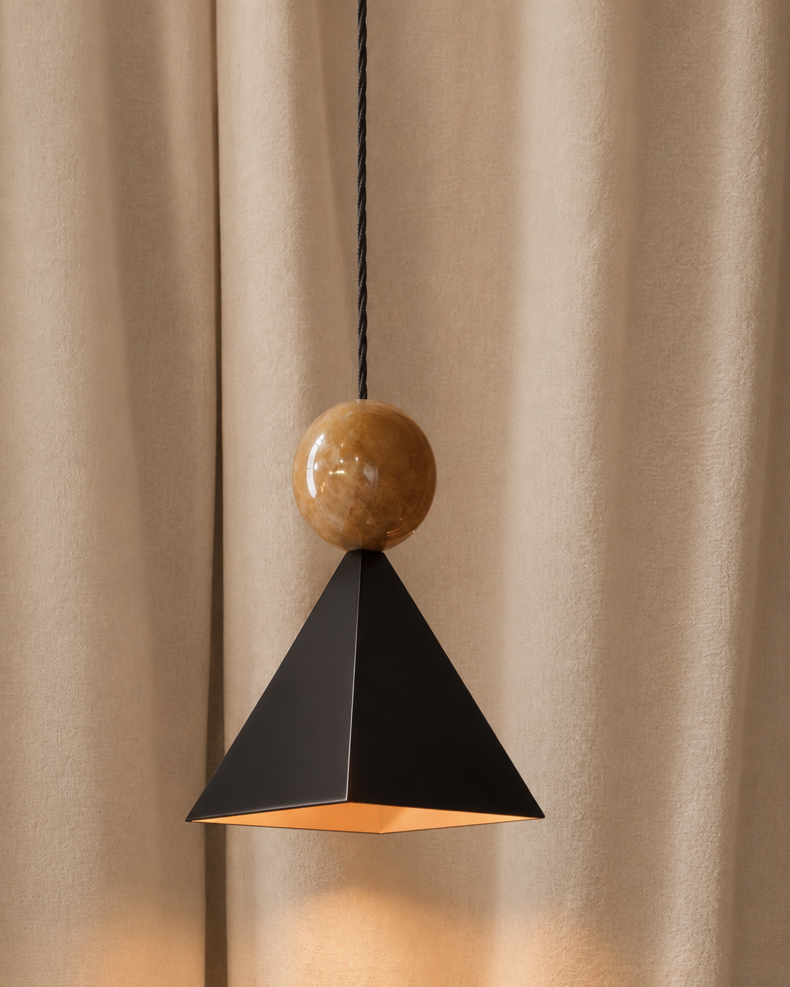 Modern pendant light with a black geometric shade and a bronze sphere, hanging in front of beige curtains.