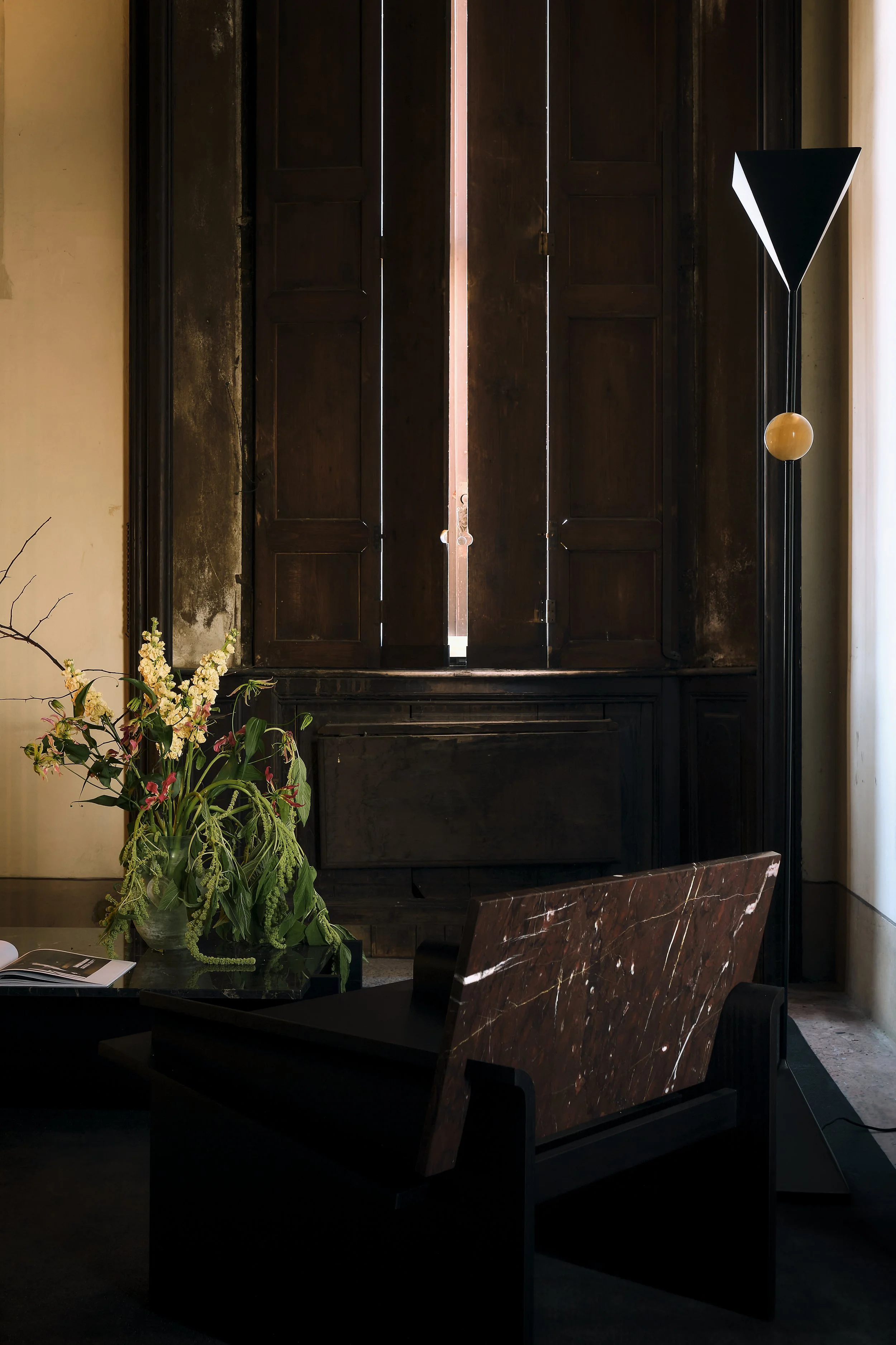 A dark wooden wardrobe with two sets of closed doors, in a room with cream-colored walls. To the right of the wardrobe, there is a modern black floor lamp with a black triangular shade and a round wooden accent. In front of the wardrobe, there is a t