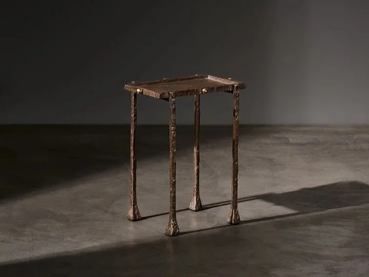 An old, rustic wooden table with four uneven, carved legs, standing alone on a concrete floor in a dimly lit room.