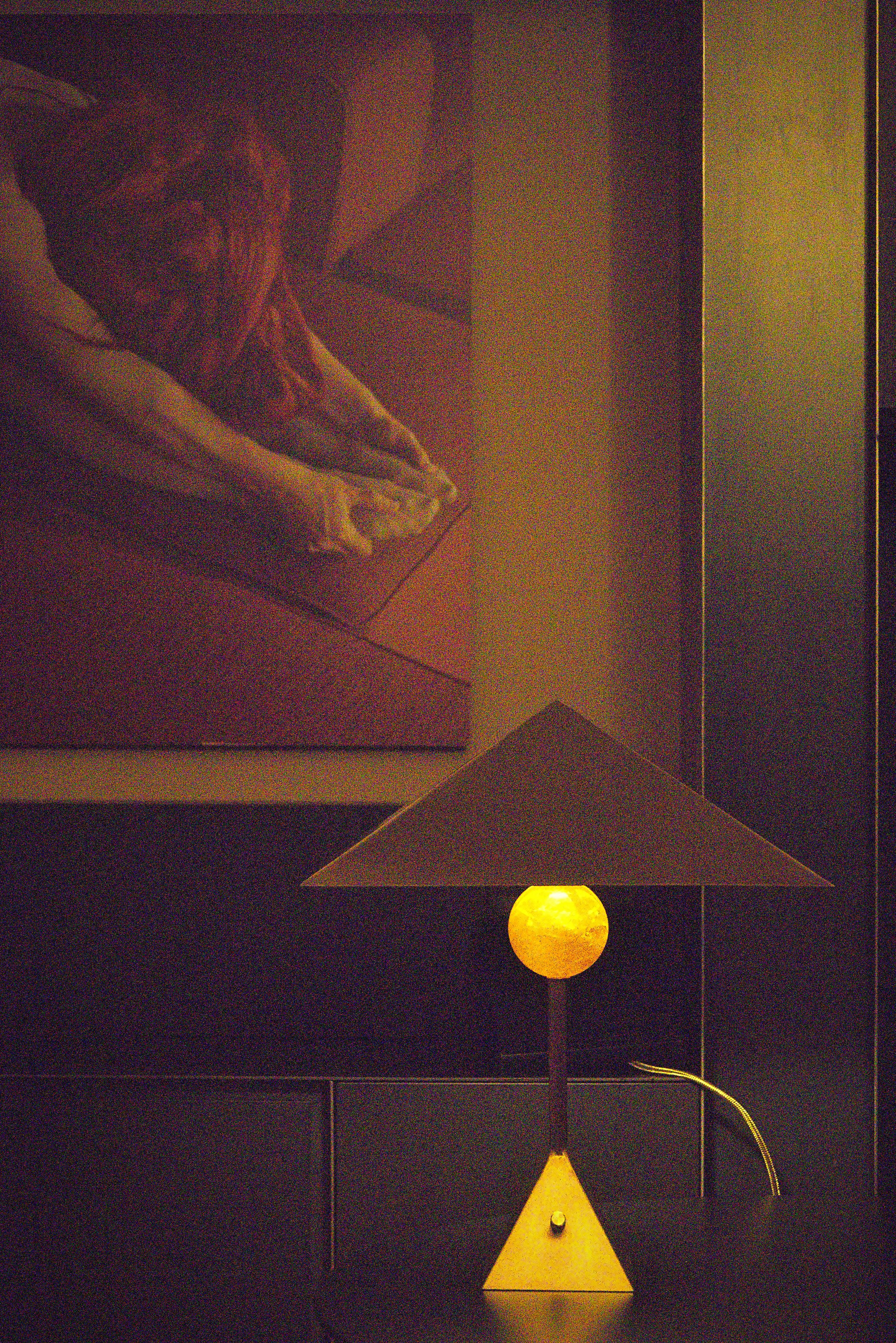 A minimalist desk lamp with a yellow spherical base, a yellow triangular shade, and a yellow vertical rod connecting them, placed on a dark surface against a dark wall background.