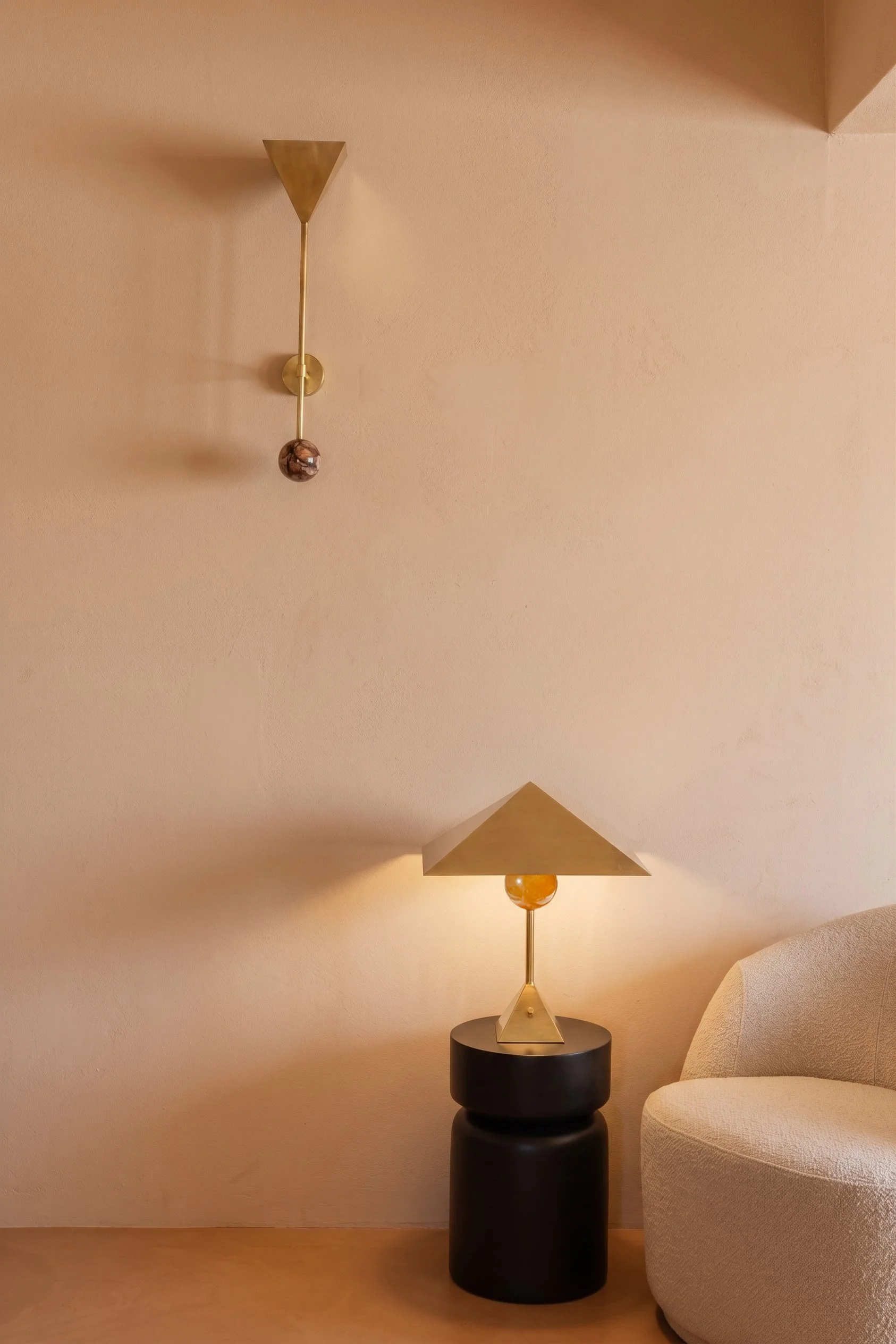 A modern table lamp with a geometric design, placed on a black cylindrical side table, next to a beige upholstered chair in a minimalist room with beige walls.