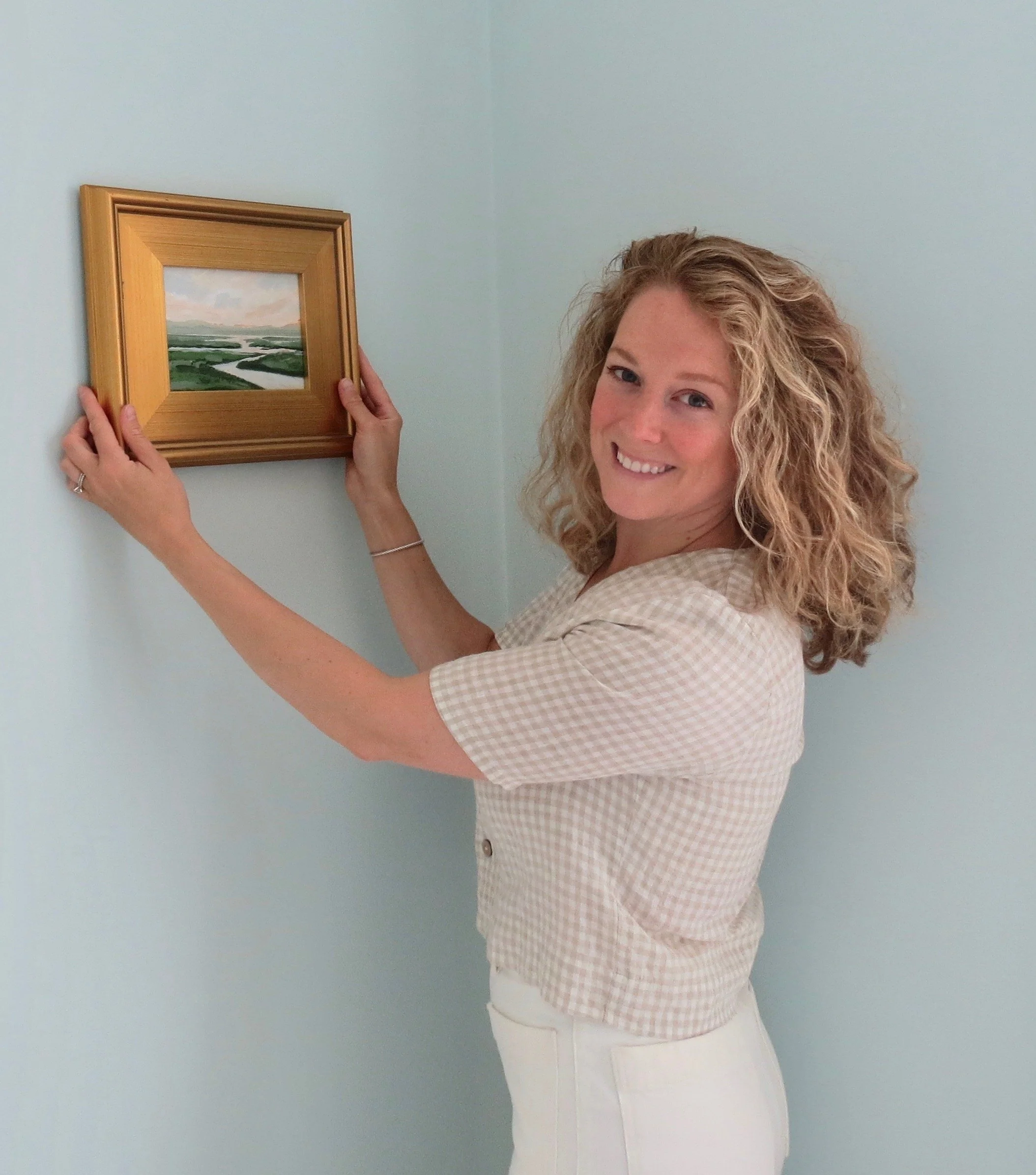 New England Artist Rachel Champa of Sea & Linen with a salt marsh original painting, framed in gold, set in front of a light blue wall, dressed in a gingham linen top