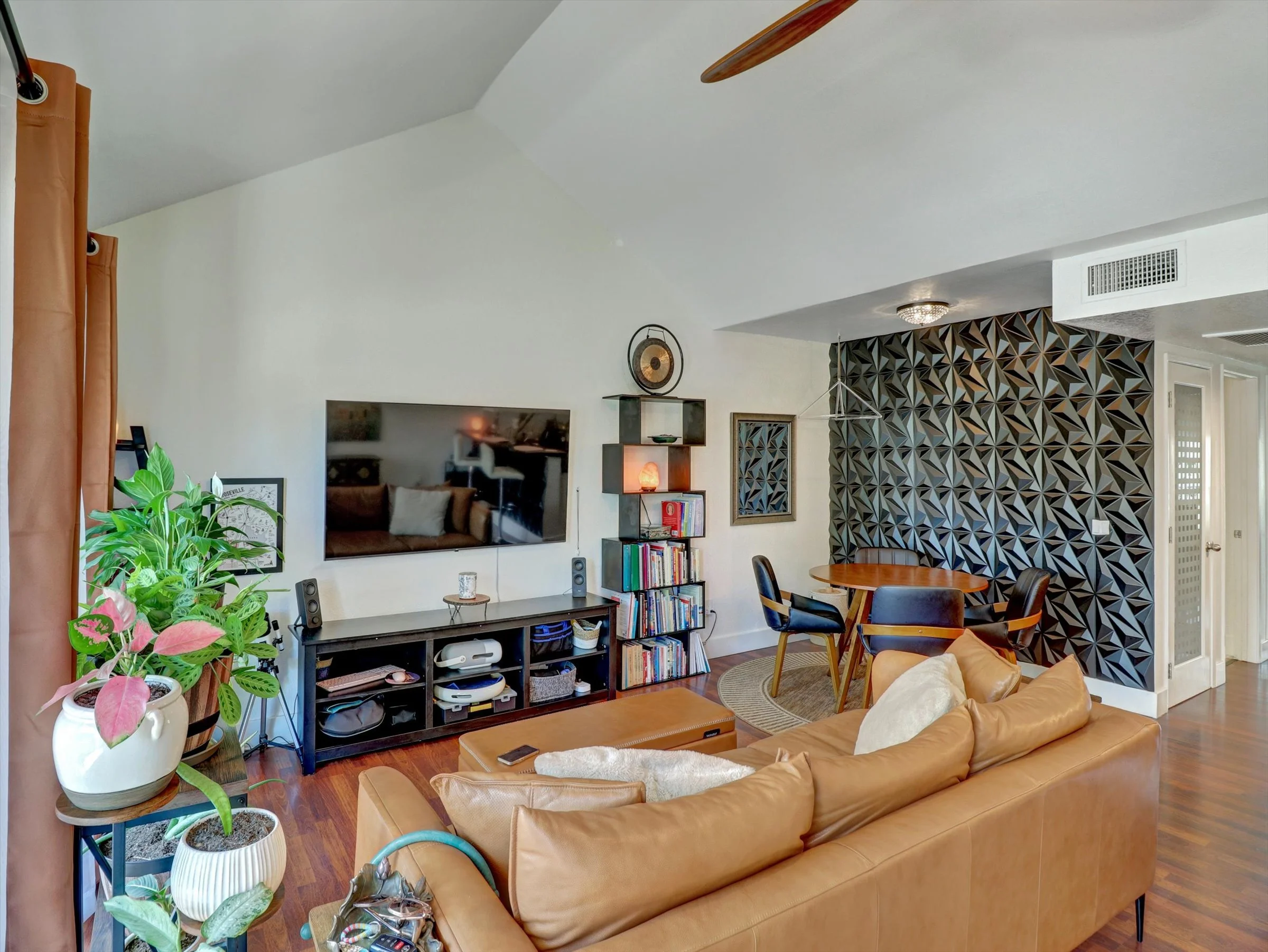 Living room with a tan leather sofa, potted plants, a wall-mounted TV, a black entertainment center with electronics, a bookcase, a dining table with four chairs, a black geometric accent wall, and hardwood floors.