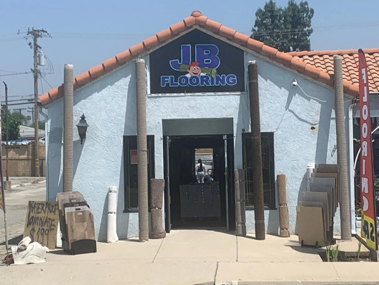 Exterior view of J B Flooring store with a sign depicting a cartoon character holding a rolled-up carpet. There are slabs of flooring materials displayed outside on either side of the entrance, and a sign on the left advertising cork flooring for $1.99. Power lines and a tree are visible in the background.