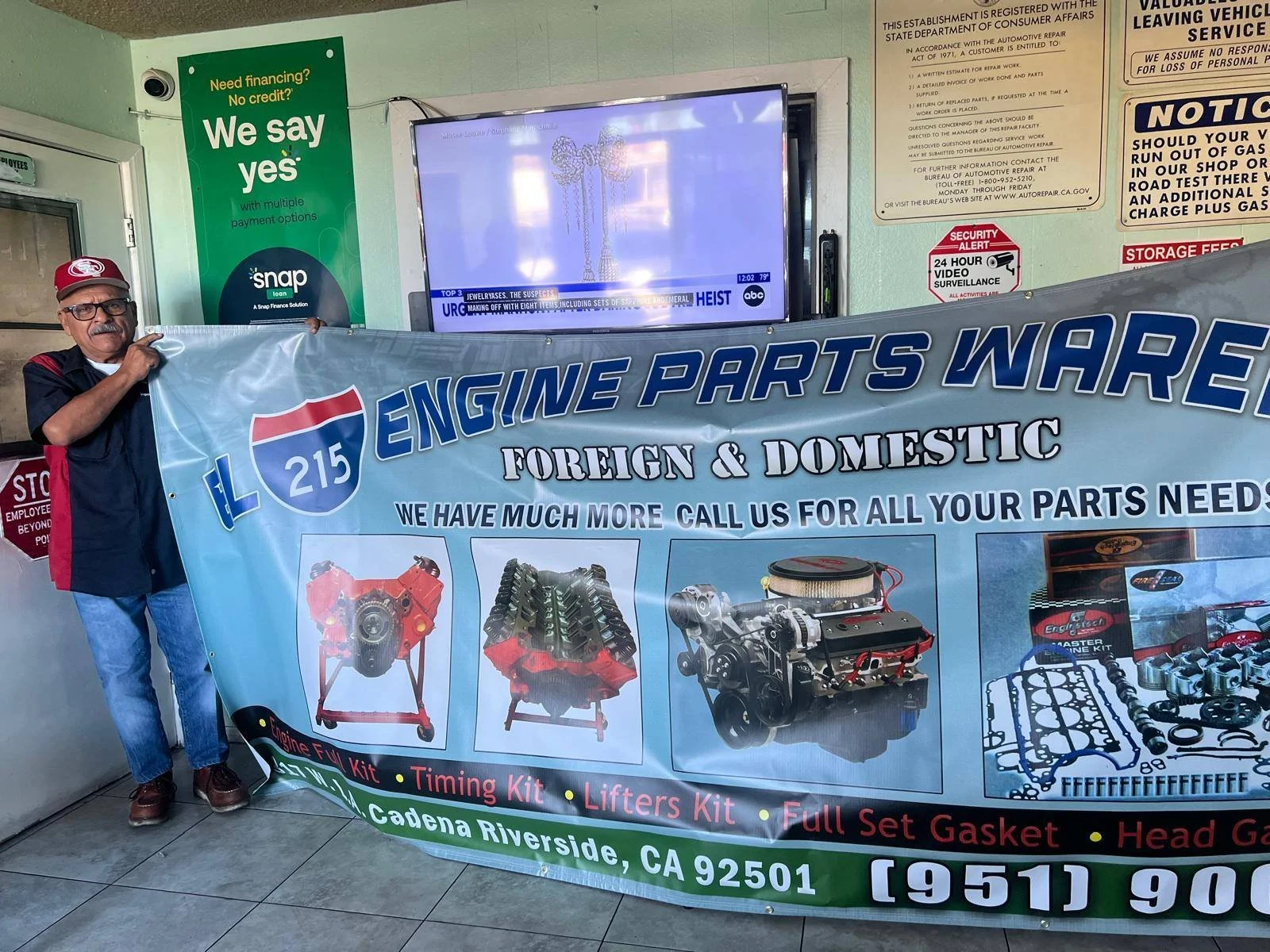 A man holding a large blue banner advertising engine parts at a store or trade show. The banner features images of engine components and text promoting the parts and contact information.