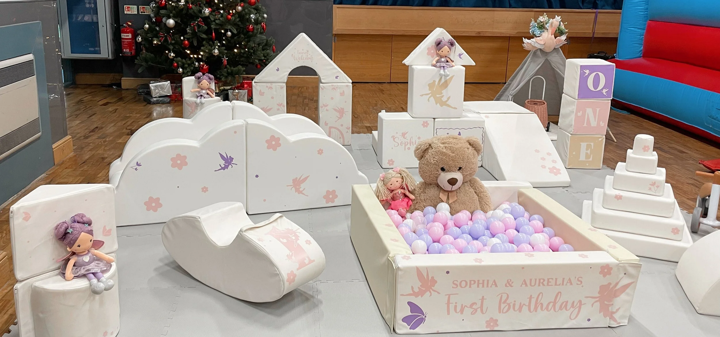 Decorative play area for a first birthday celebration with fairy-themed toys and pastel-colored balls. Includes stuffed bears, dolls, foam block structures spelling 'ONE,' and a box with the text 'SOPHIA & AURELIA'S First Birthday,' set in front of a Christmas tree.