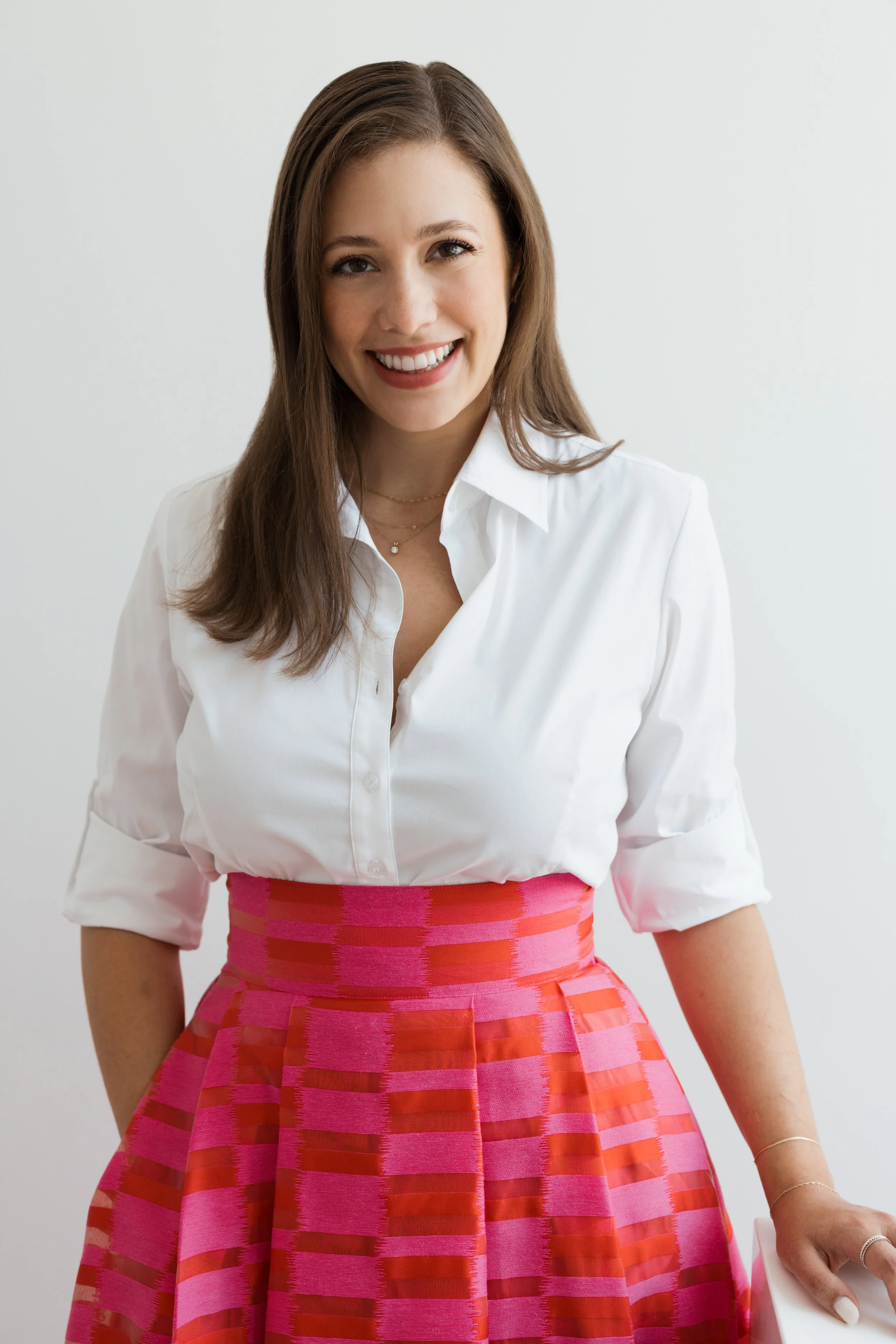 A woman with brown hair, wearing a white blouse and a pink and red plaid skirt, smiling at the camera against a plain white background.
