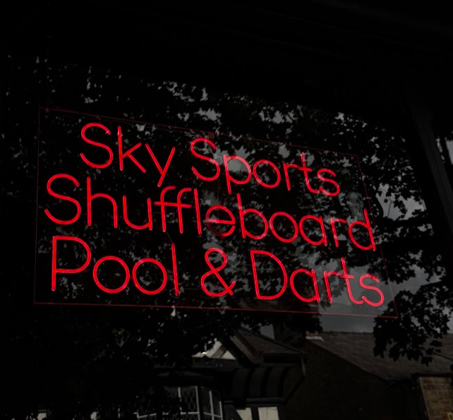 Neon sign advertising Sky Sports, Shuffleboard, Pool, and Darts in red text at Hickey's & Friends bar in Barnoldswick, Lancashire. 