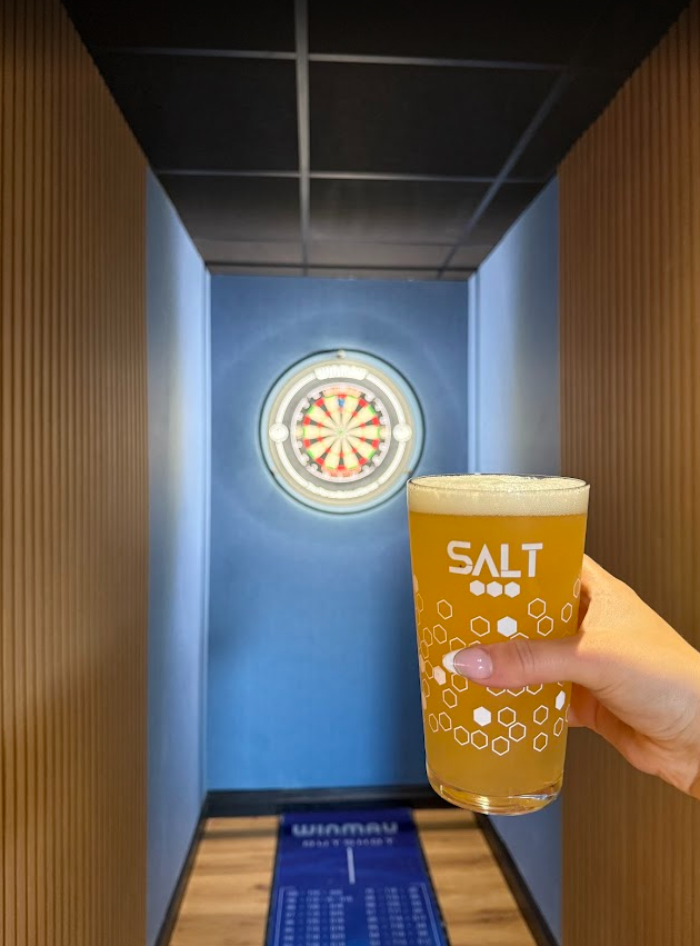 Person holding a glass of beer labeled "SALT" in front of a dartboard on a blue wall in the recreational area of Hickey's & Friends bar in Barnoldswick, Lancashire.