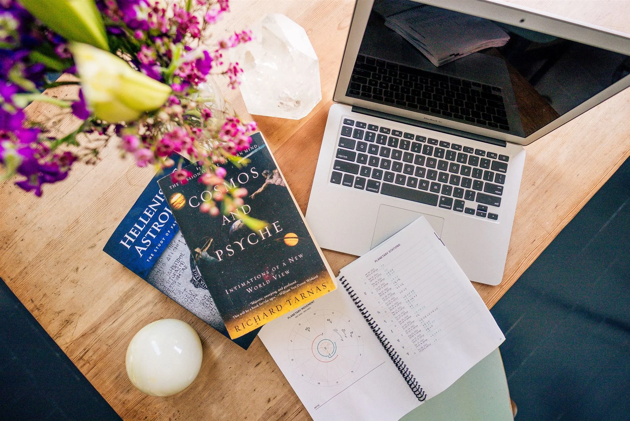 Top-down view ofworkspace featuring a laptop, two science books titled 'Hellenic and Astronauts' and 'Cosmos and Psyche', a spiral notebook with printed charts and data, a small white spherical object, a bouquet of pink and purple flowers in a jar, and a large crystal on a wooden desk.